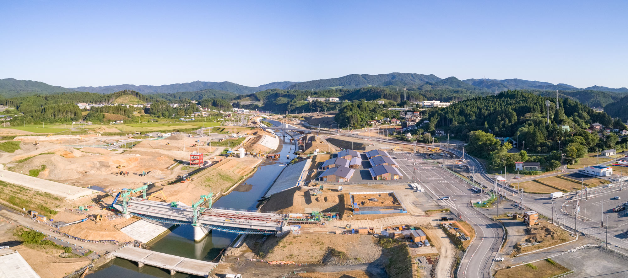 中央に川が流れ、その両岸で工事が進められている広大な建設現場を上空から撮影したもので、左側には土砂が積まれた広い土地、右側には道路や建物、さらに背景には緑豊かな山々が広がり、橋の建設も進行中である様子が写された建設現場のパノラマ写真