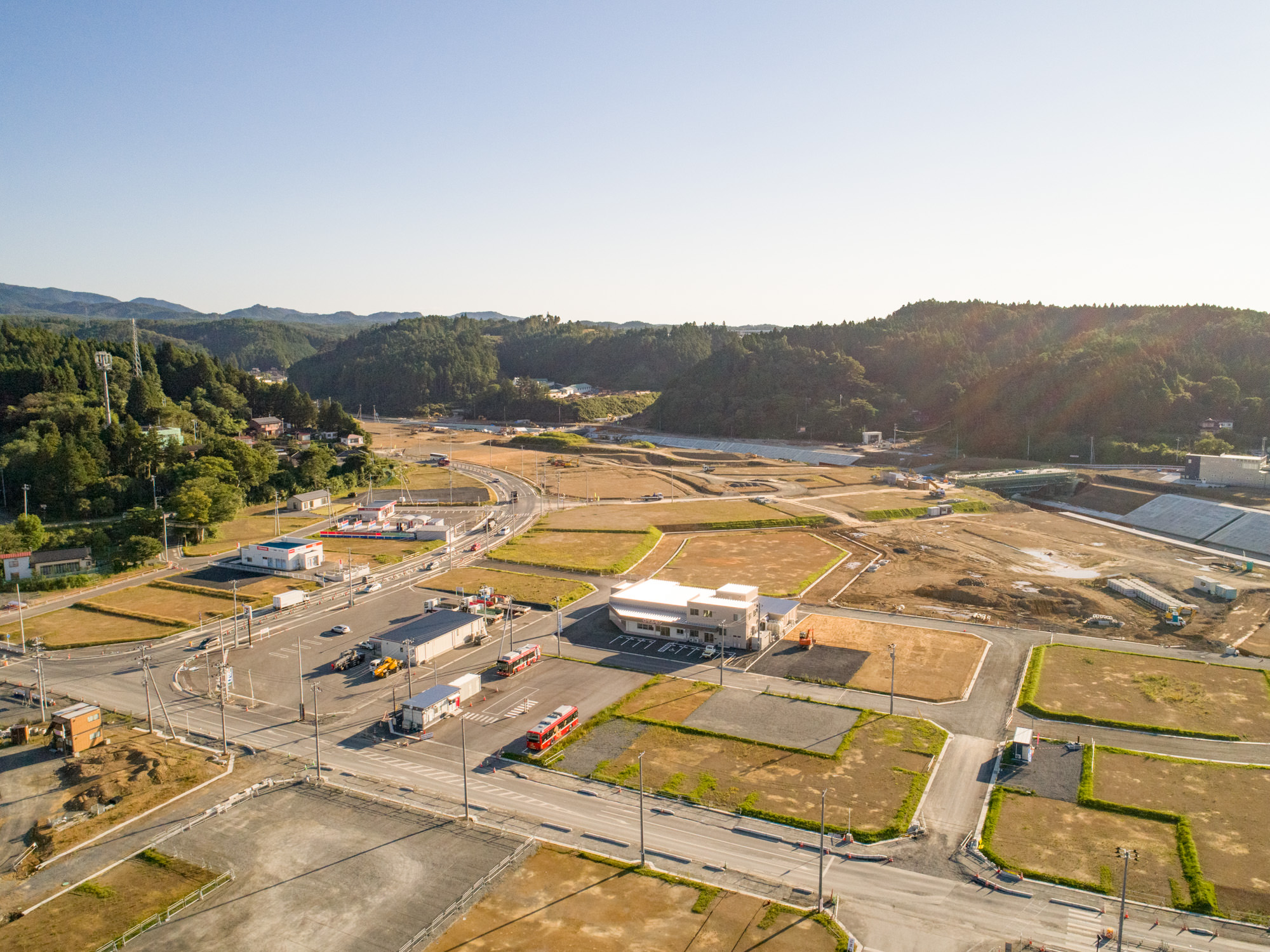 開発された土地と未開発の土地が混在する地域を上空から撮影したもので、複数の建物、道路、車両が見られ、風景には緑地や背景の丘が広がっている建設現場の写真