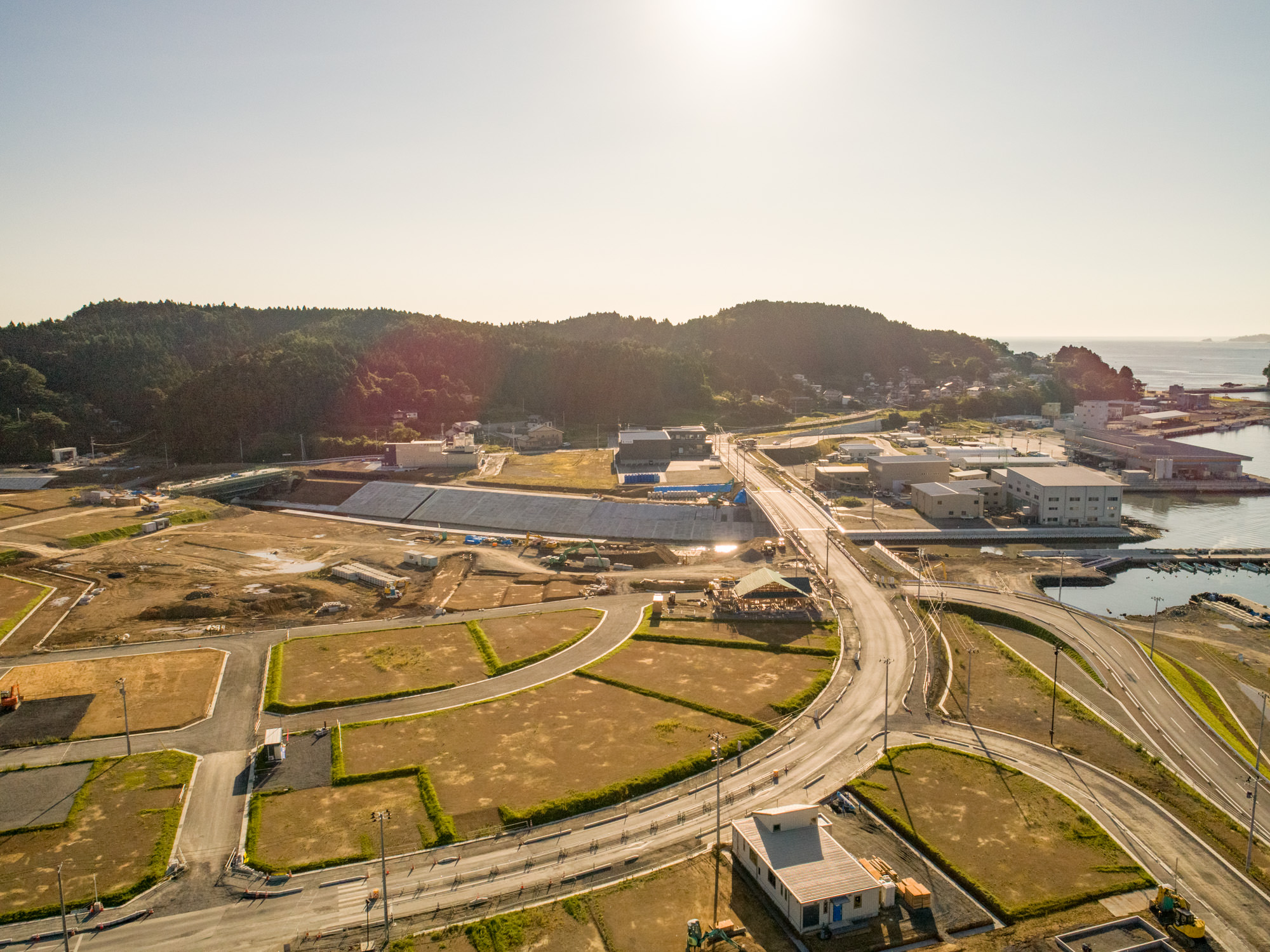 道路網といくつかの建物、工事中の土地が広がる地域を上空から撮影したもので、背景には木々に覆われた丘があり、右側には海が広がり、太陽の光が明るく差し込み、地面に影を落としている写真