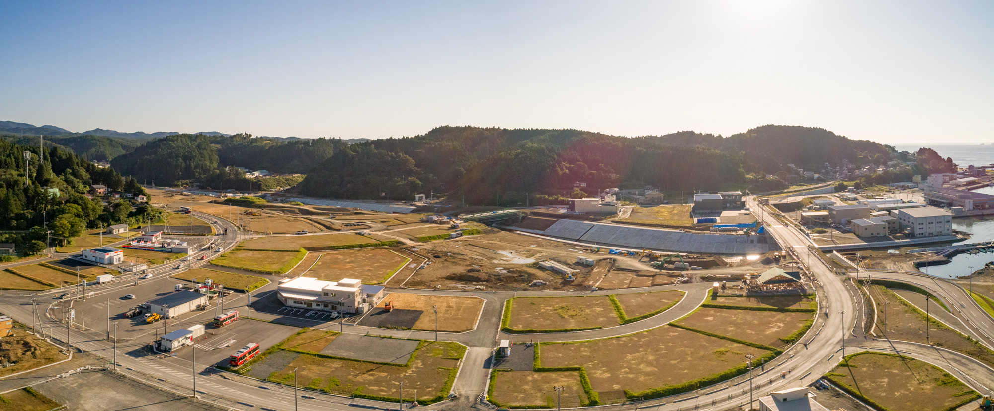 開発済みの土地と未開発の土地が混在する地域を上空から撮影したもので、手前には複数の建物、道路、駐車場があり、右側には水辺が広がり、背景には森林に覆われた丘が見え、地域全体が開発または再整備中である様子が写されたパノラマ写真