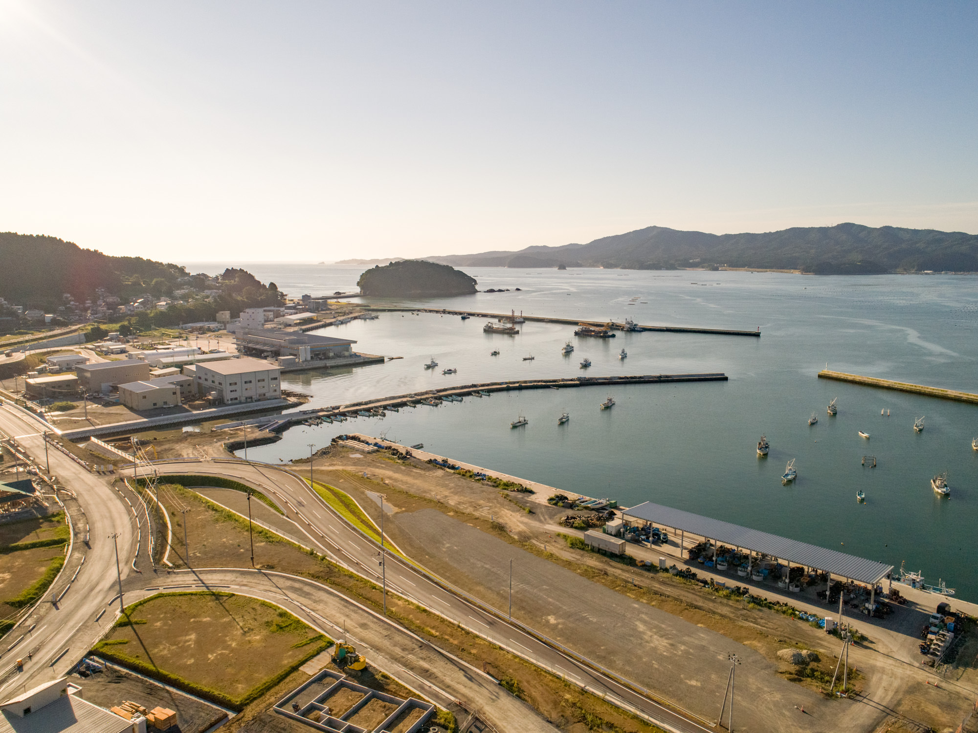 港が広がる沿岸地域を上空から撮影したもので、複数の船が水面に停泊しており、防波堤に囲まれた港の周囲には建物や道路、空き地が点在し、背景には丘や山々、さらに遠くには別の水域が広がり、全体的に穏やかで晴れた天候の建設現場の写真