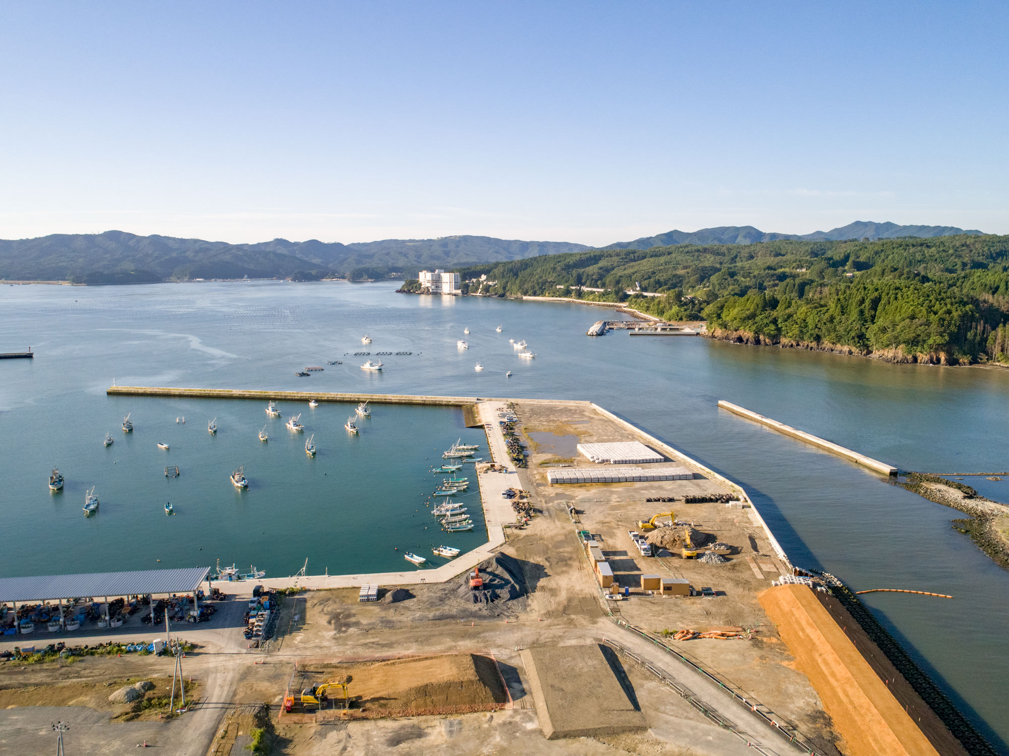 港が広がる沿岸地域を上空から撮影したもので、複数の船が水面に停泊しており、防波堤に囲まれた港の周囲には工事資材や機材が点在し、土地が開発中または整備中である様子が見られ、背景には森林に覆われた丘や山々が広がり、空は雲ひとつない晴天の建設現場の写真