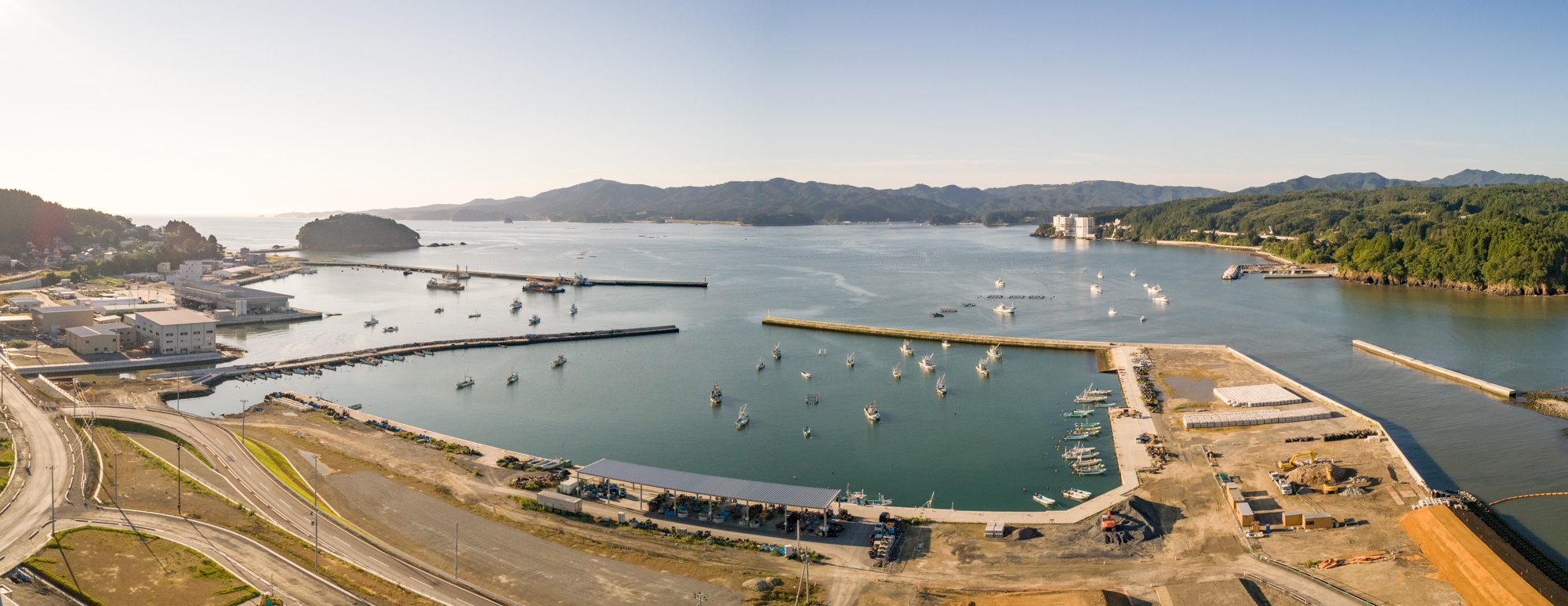 港が広がる沿岸地域を上空から撮影したもので、複数の船が水面に停泊しており、防波堤に囲まれた港の周囲には建物や道路、工業施設と思われるインフラがあり、背景には丘や山々が広がり、空は澄み渡っているパノラマ写真