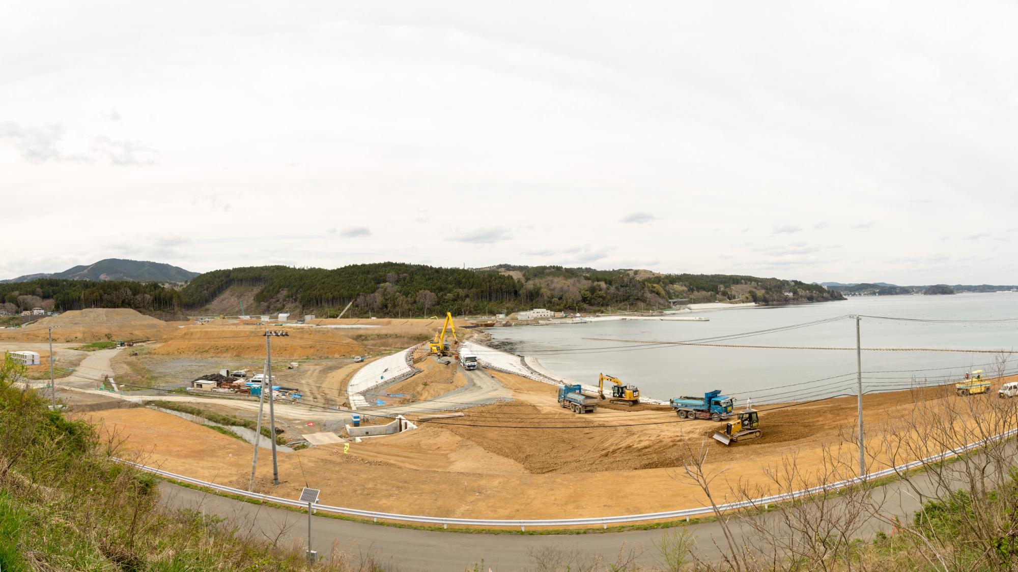 右側が海岸になっている建設現場に青いダンプやショベルカーを使い土を掘削したり移動したりしている工事の写真