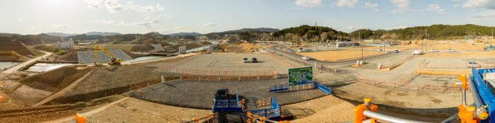 中央はフェンス囲われ、左には黄色のショベルカーがある広々とした建設現場の写真