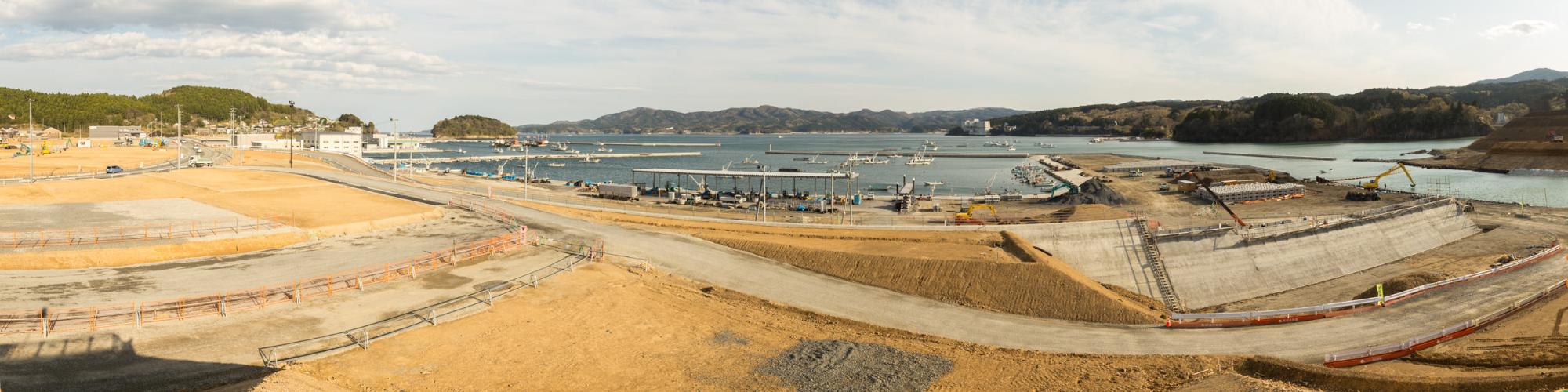 画面の手前には造成中と思われる道路が通っており、その周囲には土が盛られたエリアや工事用の柵が設置され奥には港が広がっており、多くの船やボートが停泊している様子を写した写真
