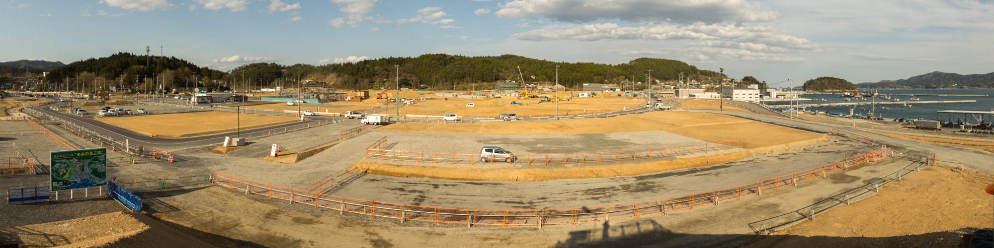 きれいに整地された建設現場で右手に海が見え、奥には山がみえ、複数の車両や建設機械が配置されている写真