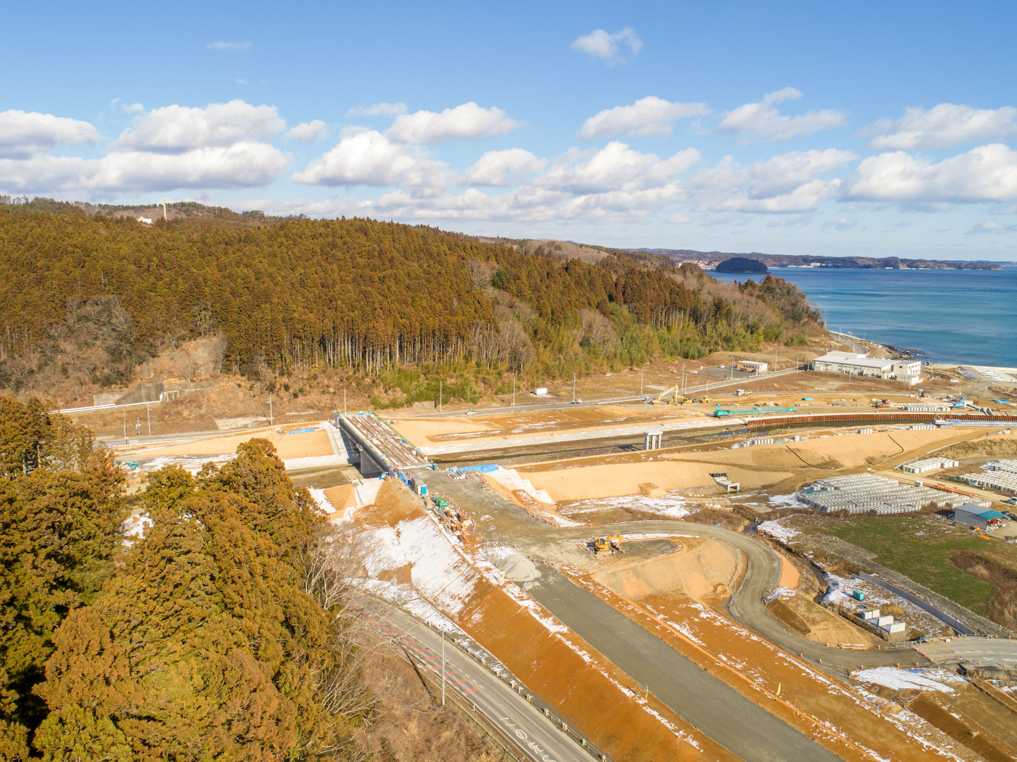 森林地帯と海岸線の近くに位置する建設現場を写したもので、複数の道路があり、一部は工事中で、重機や資材が確認でき、風景には木々と開けた土地が混在し、背景には海が広がっている建設現場の写真