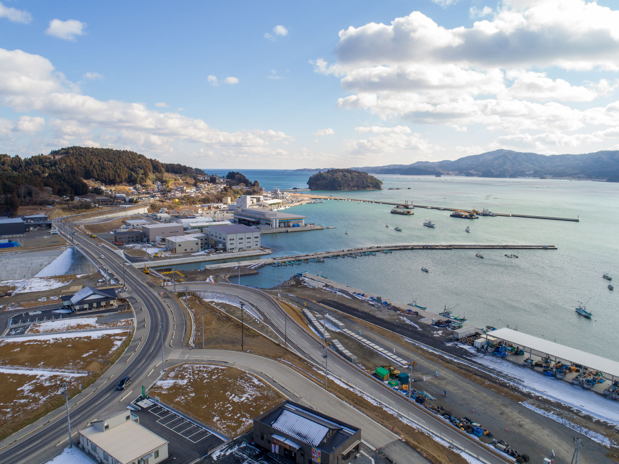 地面には雪が残っている港を中心とした海沿いの町を上空から撮影したもので、町には複数の建物や道路、海に突き出た防波堤があり、港には船が停泊していて、背景には丘や山々が広がりっている冬景色の写真