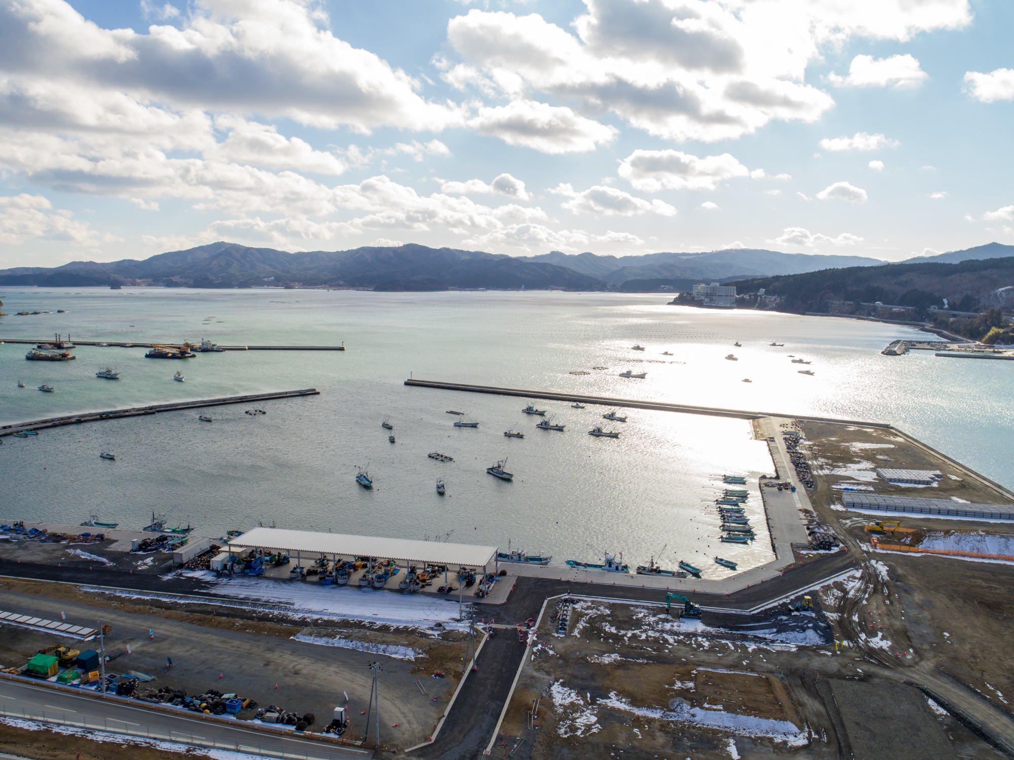 地面には雪が残っている港を中心とした海沿いの地域を上空から撮影したもので、複数の船が水面に停泊しており、港は防波堤に囲まれ、周囲の陸地には建物や構造物が点在し、背景には山々が広がっている防波堤に囲まれた港と周辺風景の写真
