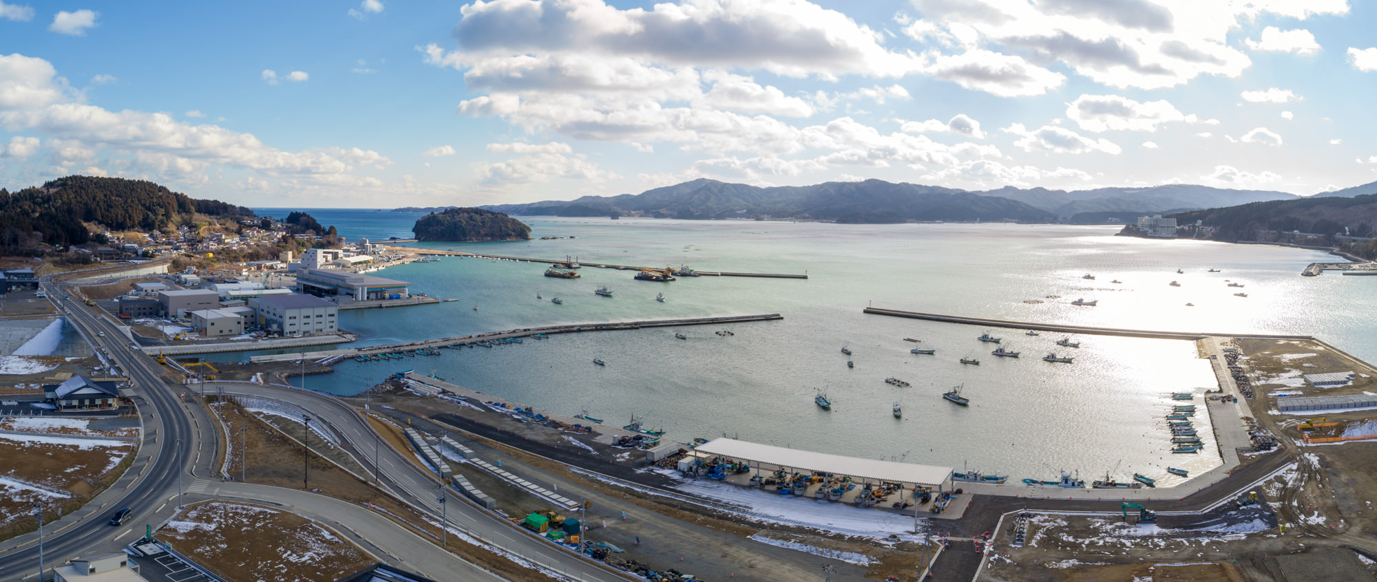 地面には雪が残っており、複数の船が停泊する海沿いの港湾地域を広く見渡したもので、港は三方を陸地に囲まれ、岸沿いには建物やインフラが整備されており、港へと続く道路が確認でき、背景には山々と雲が点在する空が広がっている港湾地域と周辺風景のパノラマ写真