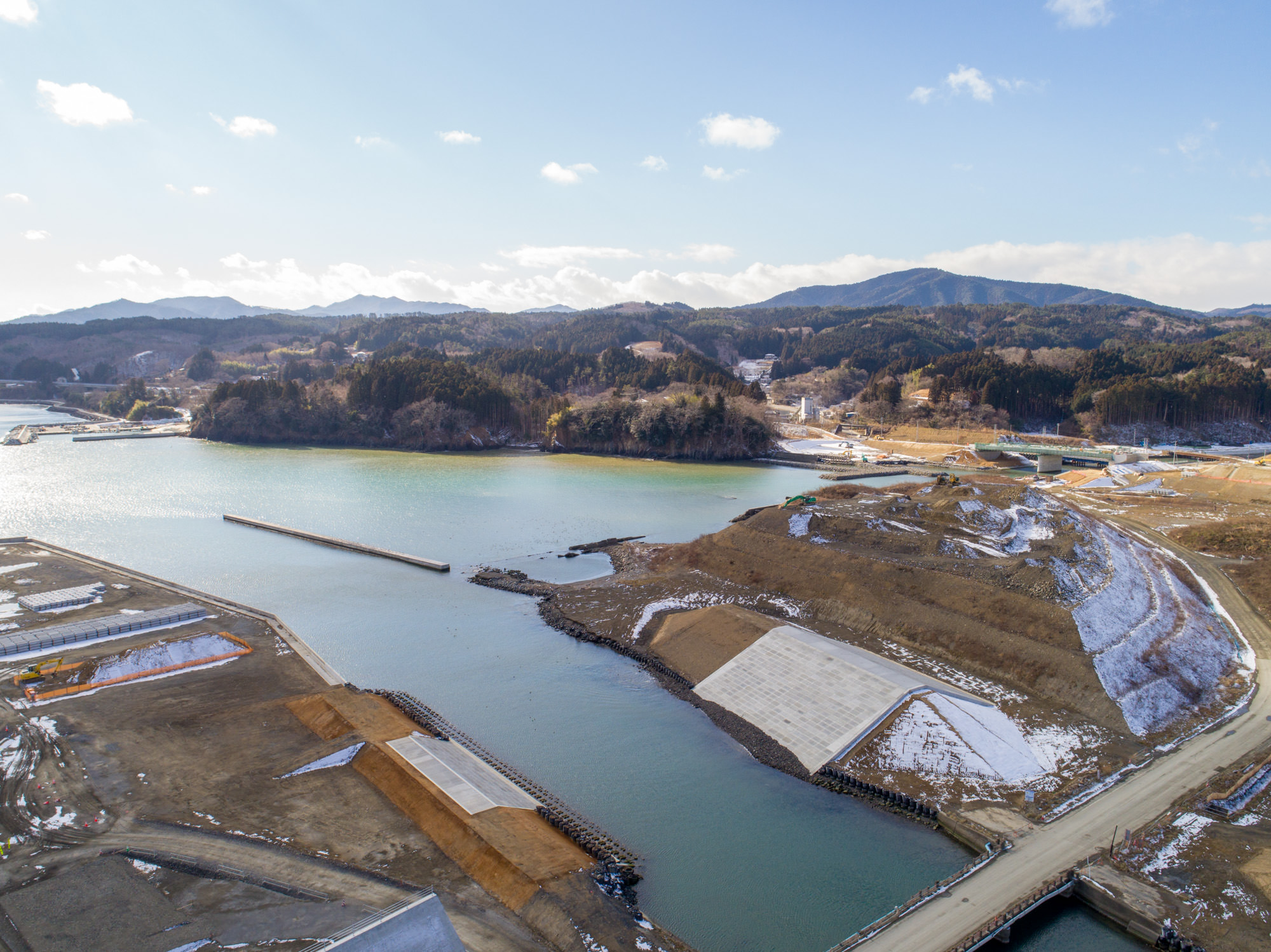 地面には雪が残っている湾や入り江と思われる水域を中心に、周囲の陸地にコンクリートの護岸や建設現場などの人工構造物が広がる海沿いの地域を上空から撮影したもので、背景には森林に覆われた丘や山々が広がっている海岸沿いの建設風景と自然地形の写真