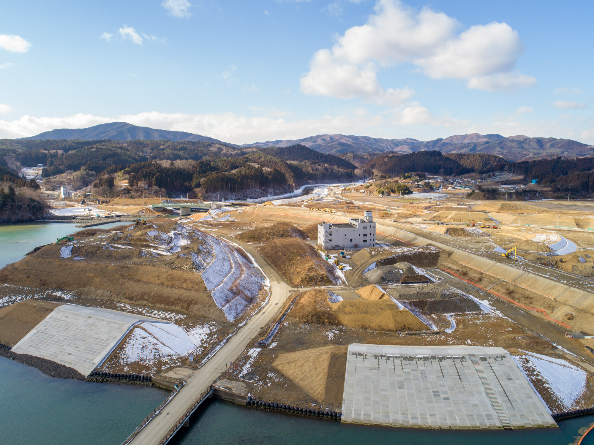 地面には雪が残る部分と地肌が見える部分が混在している自然と人工構造物が混在する風景を上空から撮影したもので、手前には水域が広がり、その両側には大きなコンクリート構造物があり、中央には奥へと延びている道路、、中ほどには複数階建ての建物があり、周囲には建設機械や資材が点在し、背景には木々に覆われた丘や山々が広がっている自然と建設が融合した風景の写真