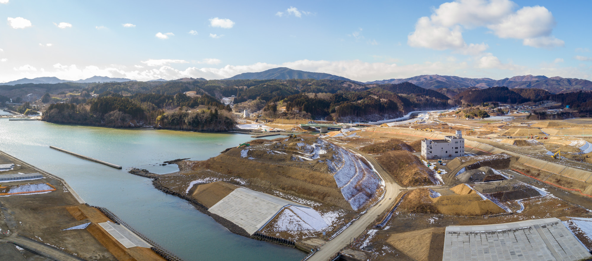 地面には雪が散在している左側に水域を望む地方の風景を広く見渡したもので、背景には丘や山々が広がり、地形には道路や構造物が点在し、中央右付近には建設途中のような建物が確認できる建設風景のパノラマ写真