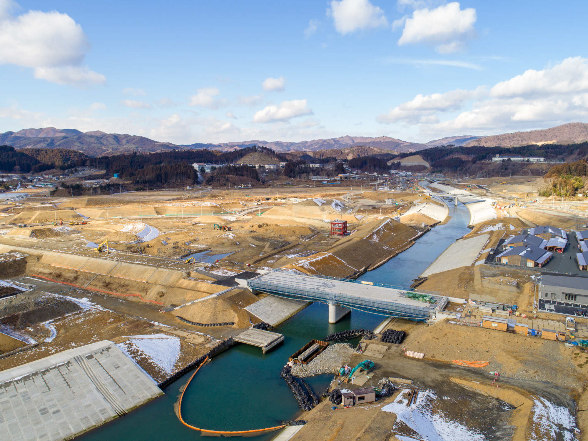 周囲は丘陵地帯で地面には雪が残っている川が流れる建設現場を写したもので、複数の橋が川を横断しており、現場にはさまざまな建設資材や機械が点在し、建設現場の近くには建物や構造物も確認できる川と橋を含む建設現場の写真