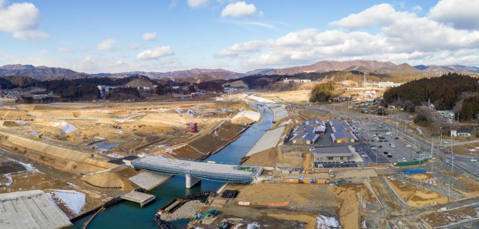 中央には川が流れ、その上には新しく建設された橋が架かっており、背景には丘や山々が広がり、地面には雪がまだらに残っている様子が見られ、右側には複数の建物や駐車場があり、開発が進んでいることがうかがえる復興の様子を写した写真