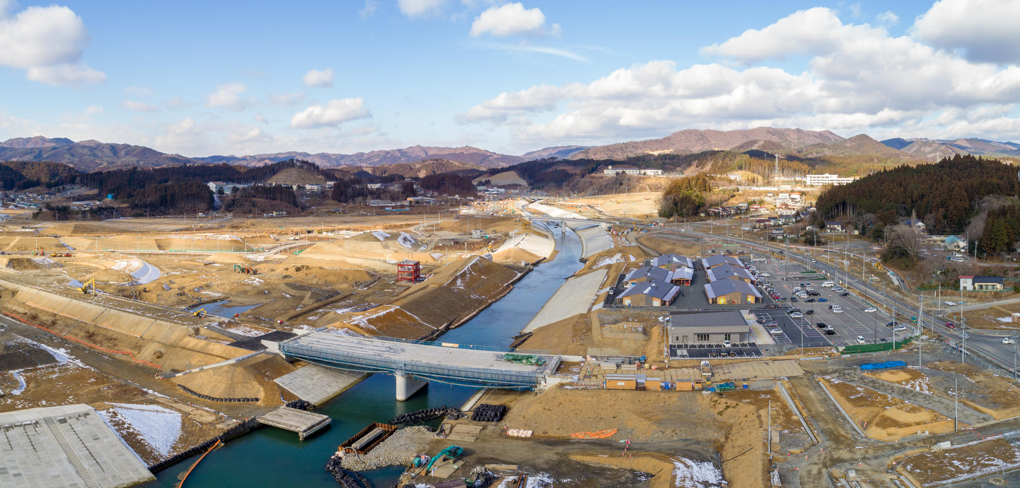 志津川地区の建設現場を広く見渡したもので、中央には川が流れ、複数の橋が架かっており、地面はほとんどが開けた土地で一部に雪が残っている冬の季節に建設資材や機械が点在し、背景には丘や山、森林が広がり、右側には駐車場と複数の建物が確認できる川沿いの地方建設風景の写真