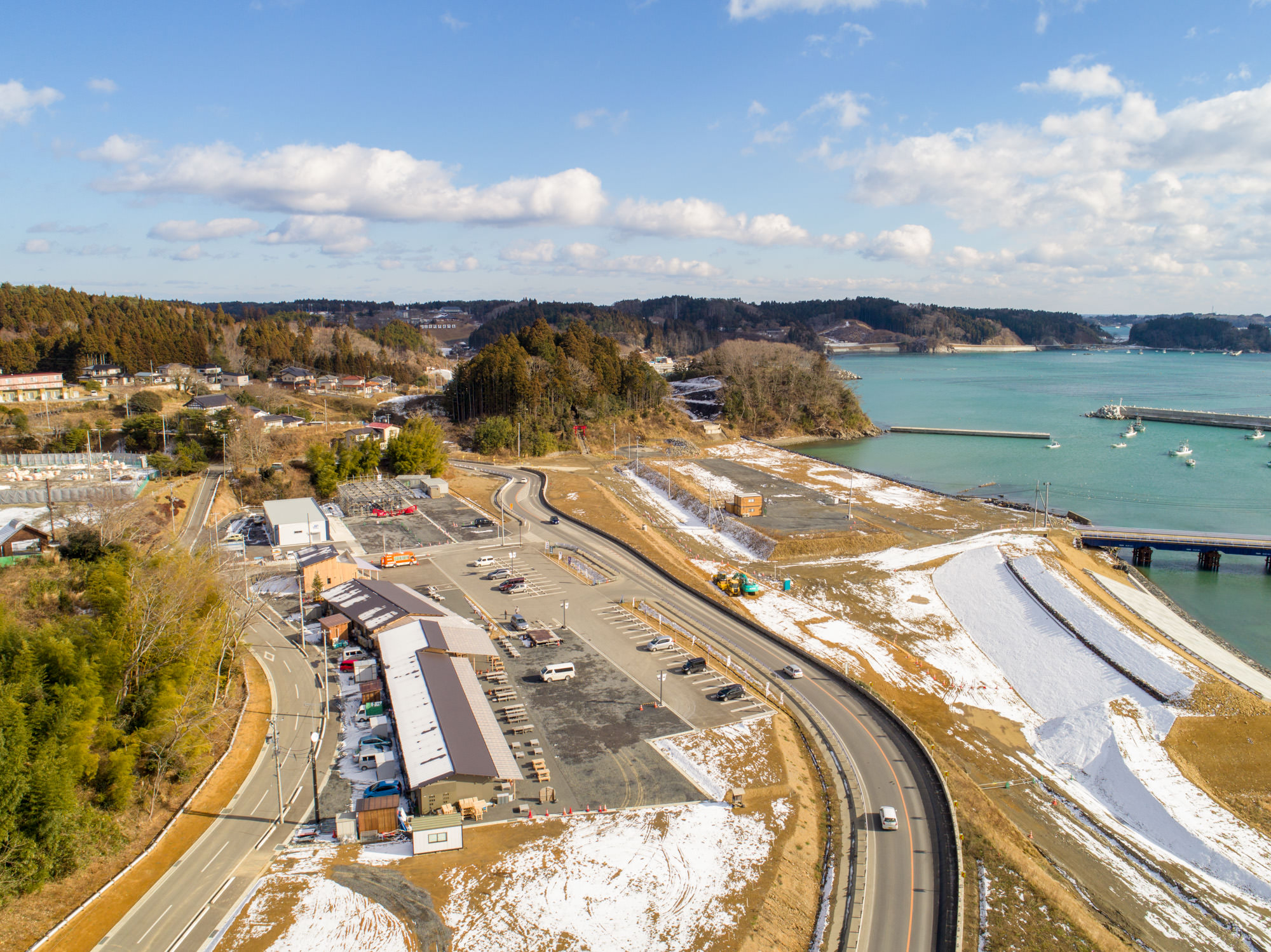雪が部分的に積もる寒冷地の海沿いを空から撮影したもので、中央には車が停まる広い駐車場と建物があり、周囲には道路が走り、背景には木々に覆われた丘と船が停泊する桟橋のある水辺が広がる風景写真