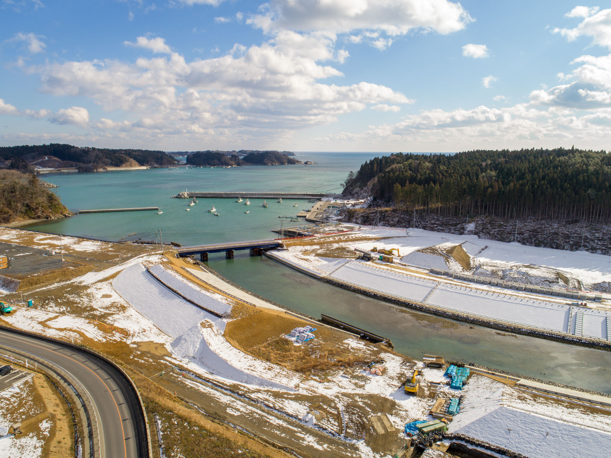 雪が積もった川沿いの地域を空から撮影したもので、中央に橋が架かる川が流れ、左側には船が停泊する湾、右側には森林地帯が広がり、手前には道路や建設中のエリアが見える晴天の風景写真