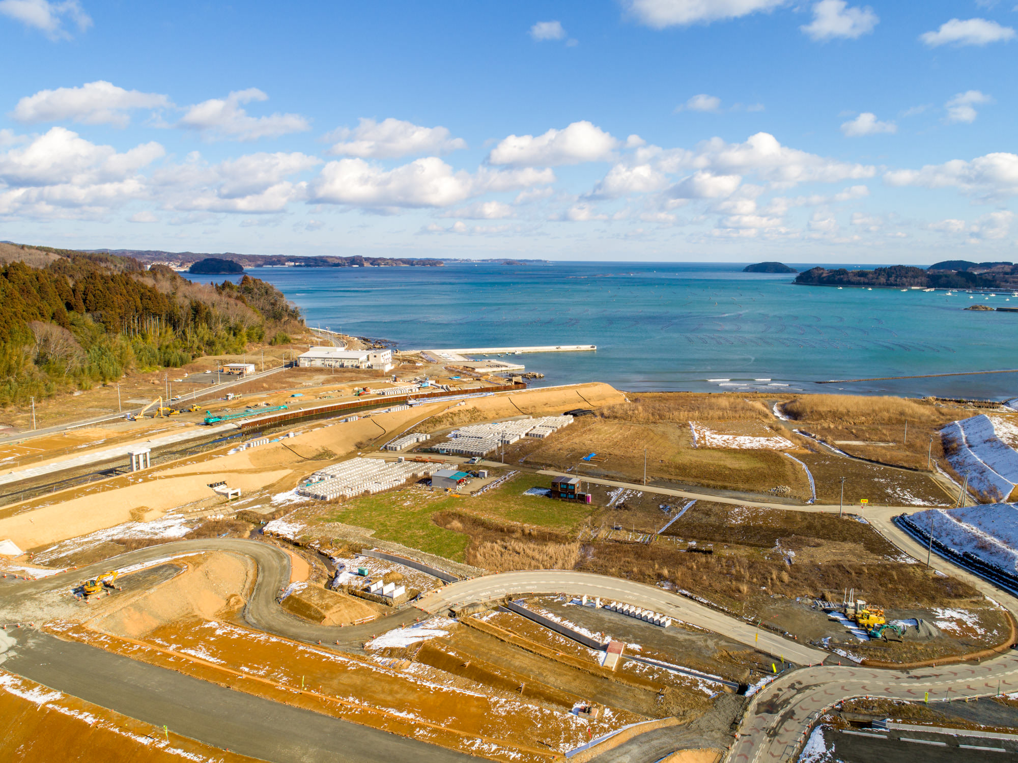 海岸沿いで進行中の建設現場を写したもので、複数の曲がりくねった道路や建設車両、建設途中の構造物が確認でき、背景には海が広がり、地面には雪が残っている海岸沿いの建設風景の写真