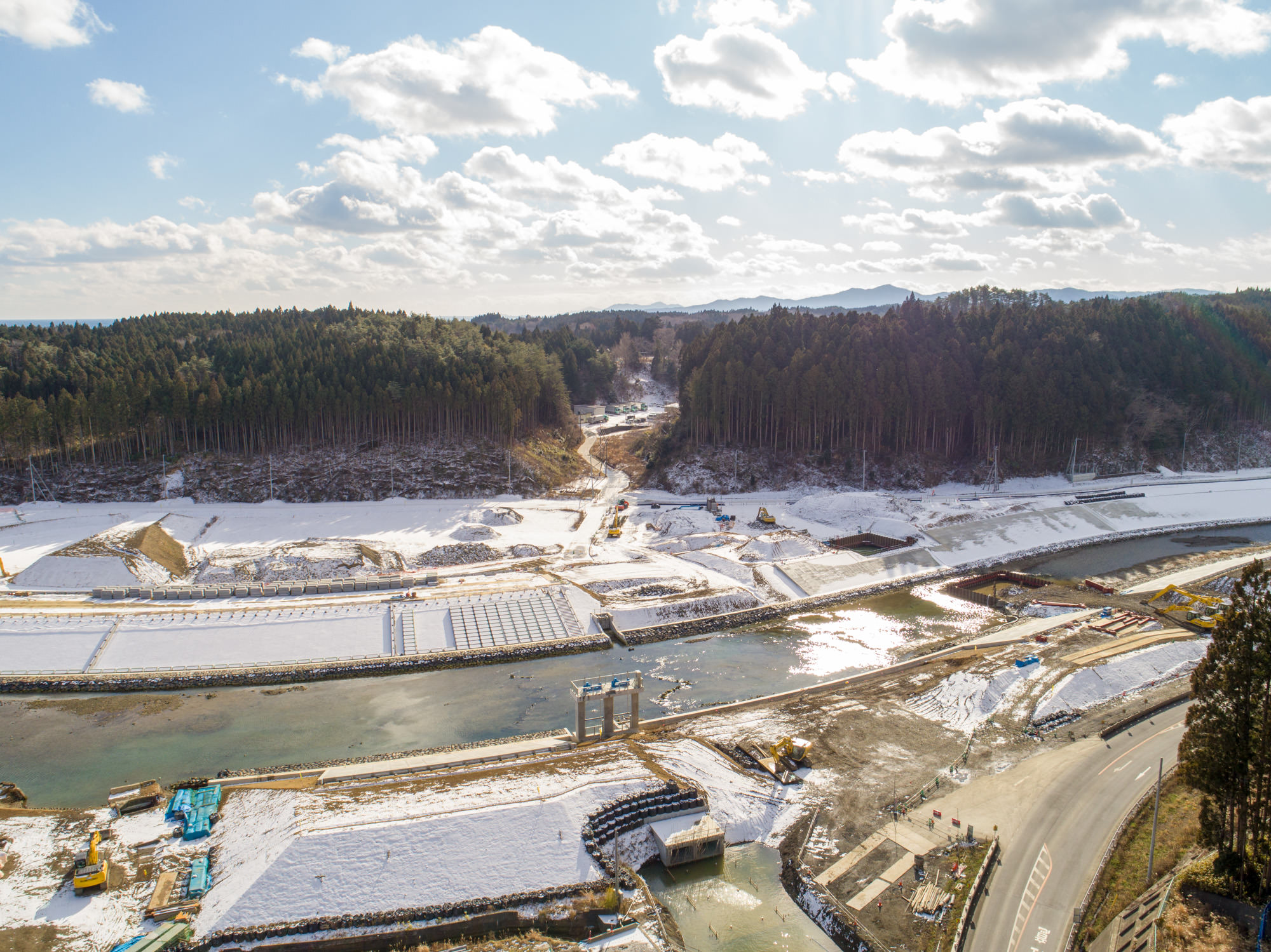 雪に覆われた風景の中にある建設現場を上空から撮影したもので、中央には川が流れ、その両岸には建設資材や機械が点在し、周囲は密集した森林に囲まれており、空には雲が点在し、差し込む日差しが一部の風景を明るく照らしている雪景色の中の川沿い建設現場の写真