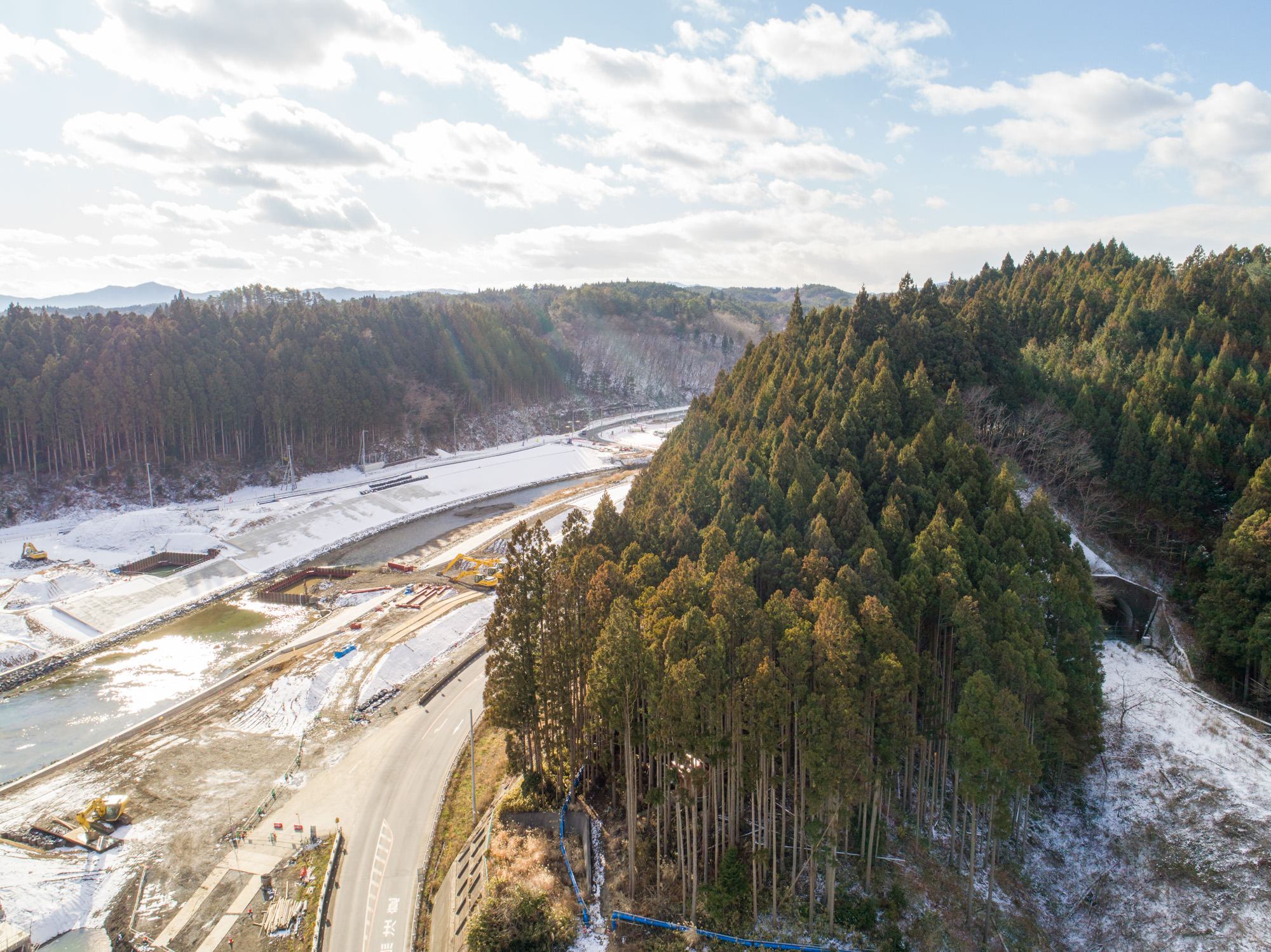 左側の川沿いには雪が積もっており、右側に背の高い木々が密集する森林が広がり、中央には川が流れる自然豊かな風景を上空から撮影したもので、森林の縁を道路が曲線を描いて通っていて、川岸付近には建設資材や機械が確認でき、空には雲が点在し、差し込む日差しが風景を明るく照らしている森林と川、建設資材が見える自然風景の写真