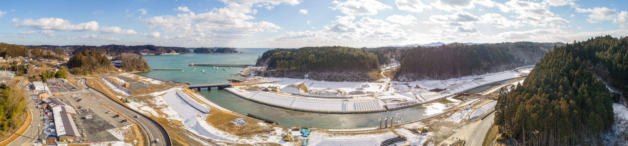 地面には雪が残っている港を中心とした海沿いの地域を広く見渡したもので、周囲には丘や森林が広がり、港には複数の船が停泊し、海岸沿いには建物や構造物が点在し、川が海へと流れ込み、その上には橋が架かっている港湾地域と自然風景のパノラマ写真
