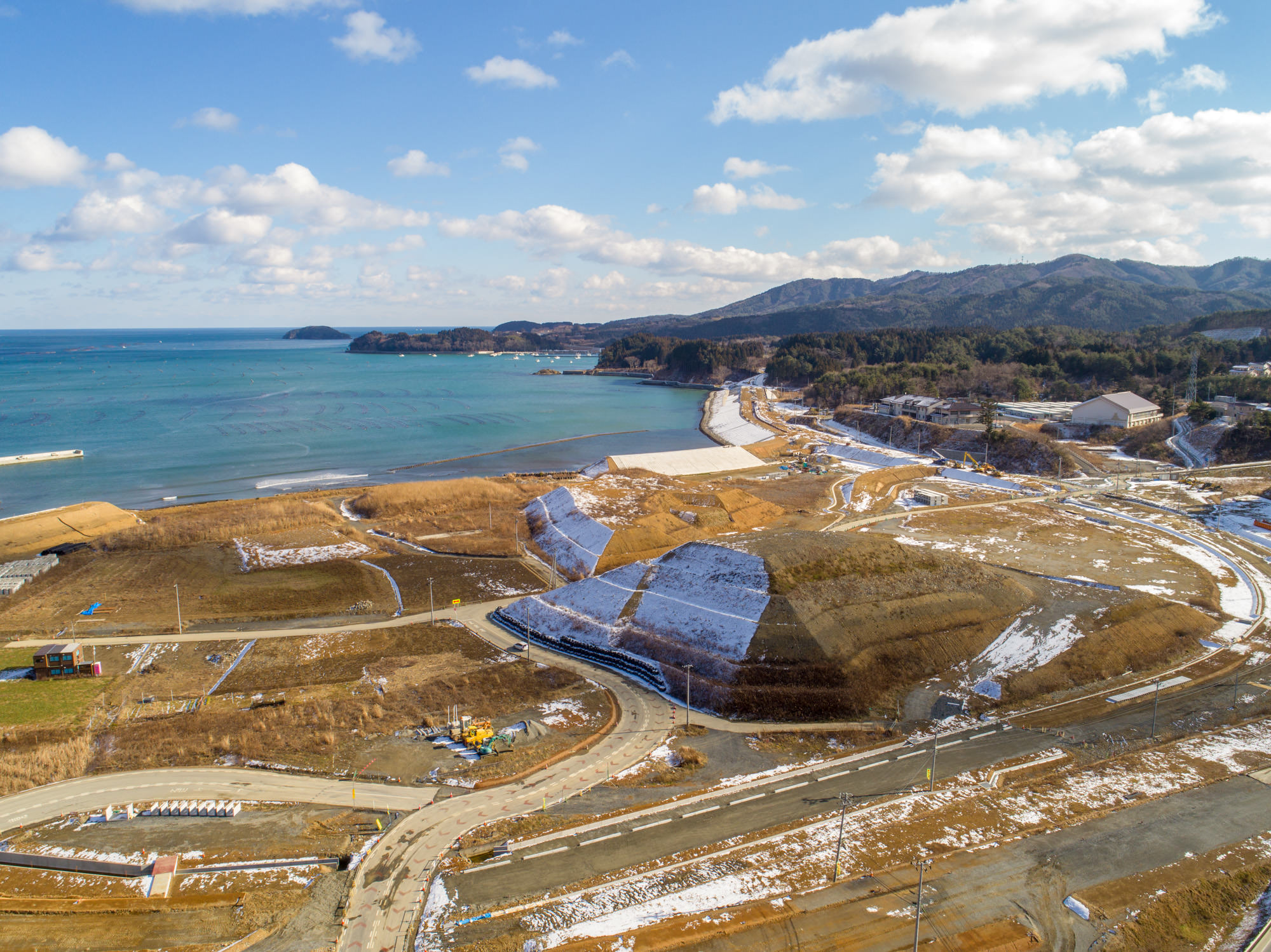自然と開発が混在する海岸の風景を写したもので、左側には海が広がり、沿岸の土地には雪が残り、道路や小道、建設機械が見られ、背景には木々に覆われた丘や山が広がっている海岸の自然と開発風景の写真