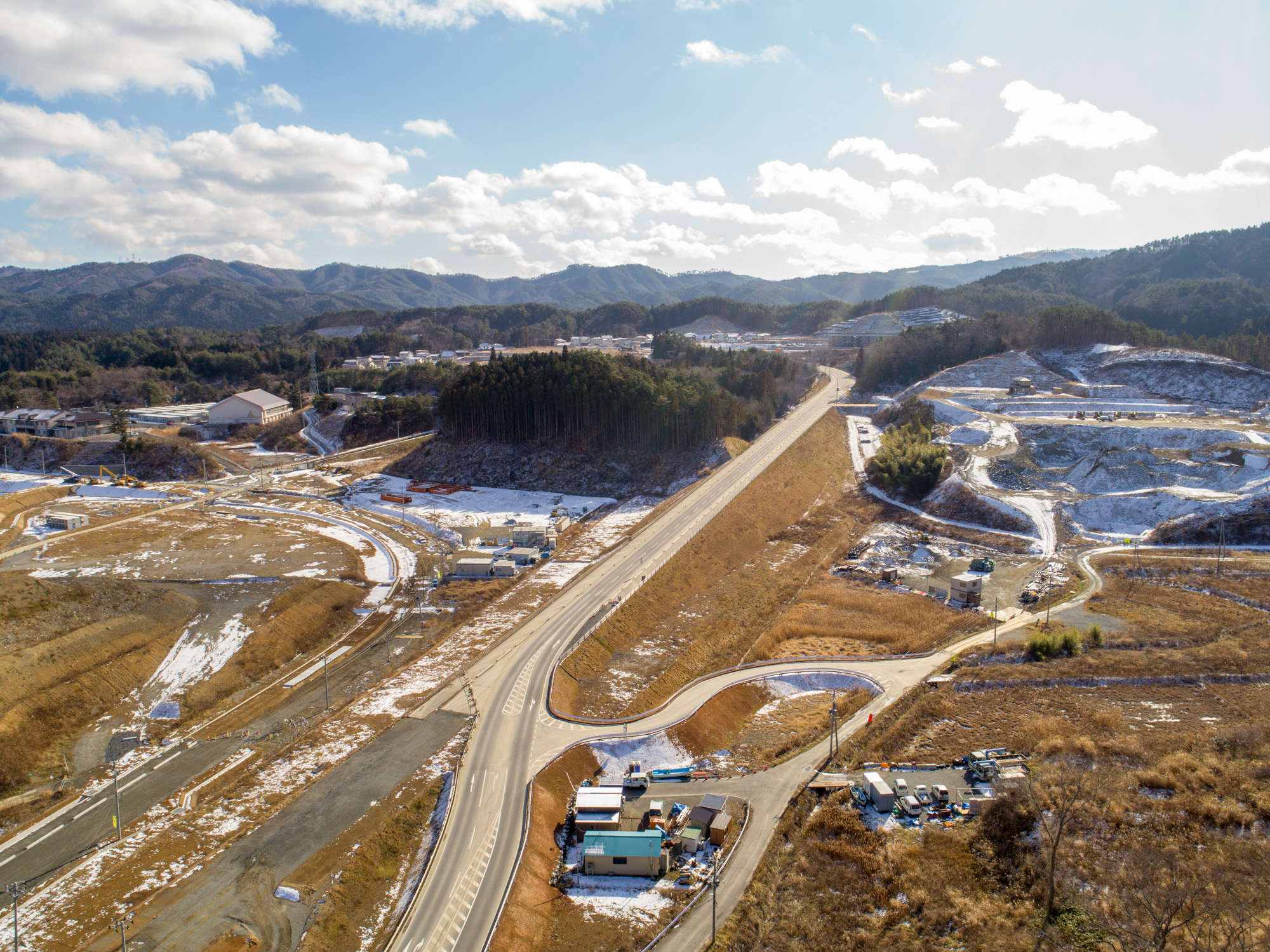 地面には雪が残っている戸倉地区の風景を上空から撮影したもので、中央には道路が通り、周囲には丘や山々が広がり、道路沿いや離れた場所に複数の建物や構造物が点在し、地形には開けた土地と植生のある区域が混在している冬の風景と建設中の道路の写真