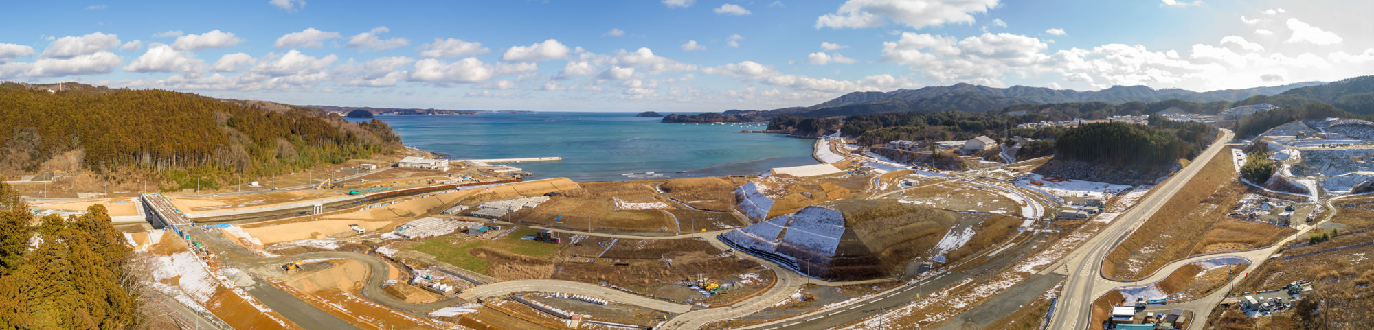 澄んだ青い海に面した海岸地域をパノラマで捉え、丘や森林に囲まれた自然と開発地が混在し、道路や建設現場、建物が点在し、地面には雪が残っている様子の写真