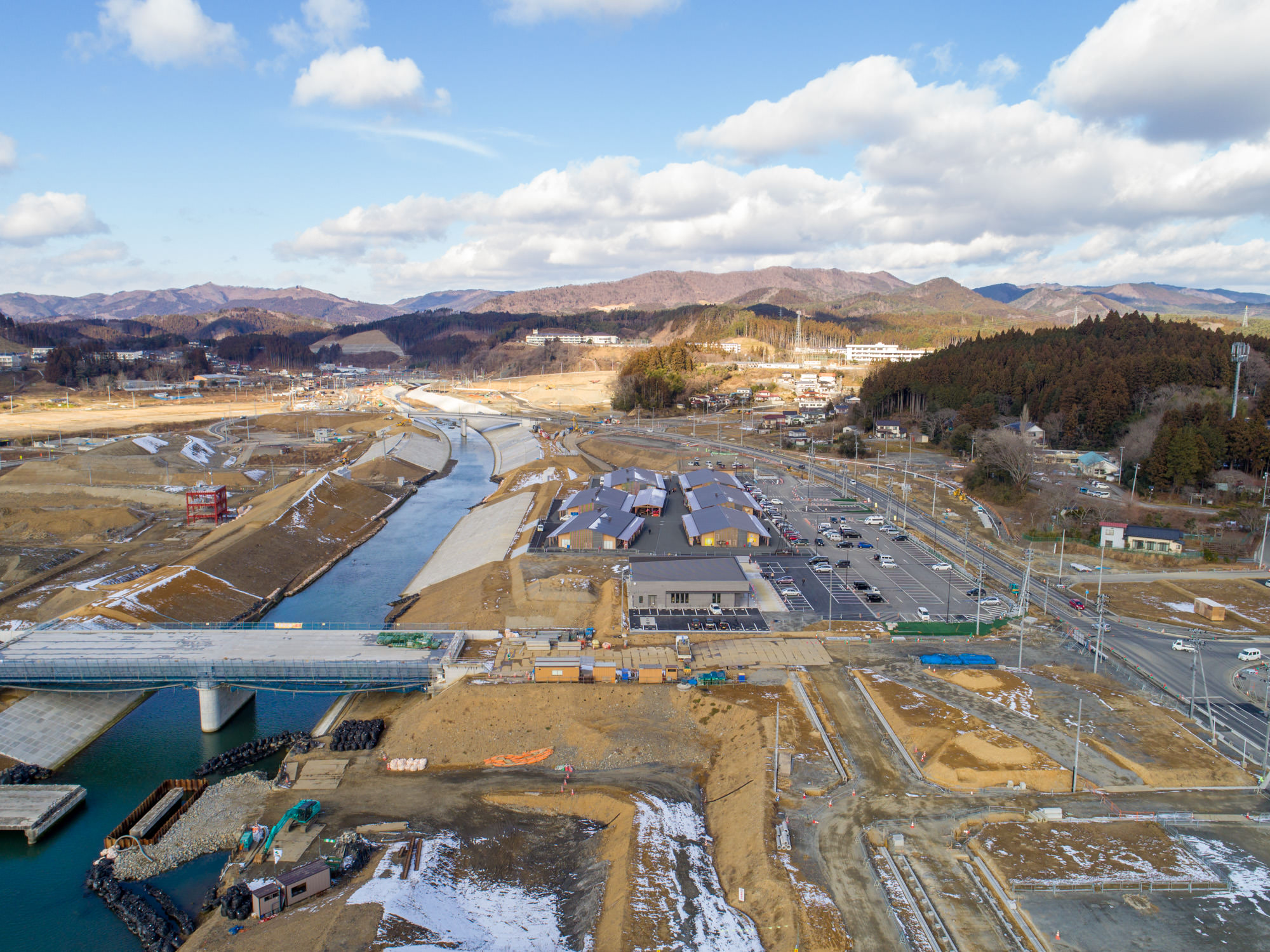 地面には雪が残っている志津川地区を上空から撮影したもので、中央には川が流れ、その上には橋が架かっており、川の両岸には複数の建物や駐車場が広がり、道路や建設現場も確認でき、背景には丘や山々が広がっている建設風景の写真