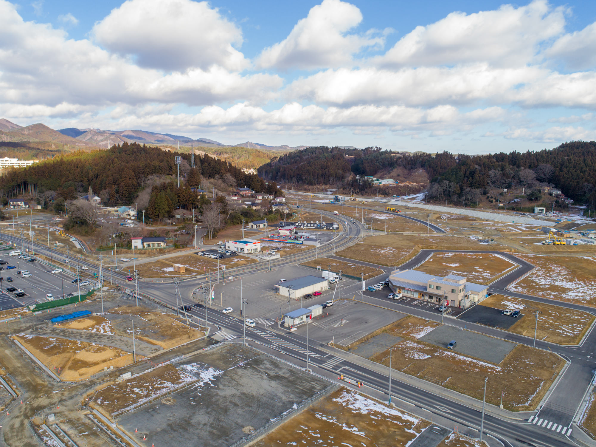 地面には雪がまだらに残っている町を上空から撮影したもので、建物や道路、広場が混在し、複数の交差点や駐車場が確認でき、周囲には森林に覆われた丘や遠くに山々が広がっている町並みと自然風景の写真