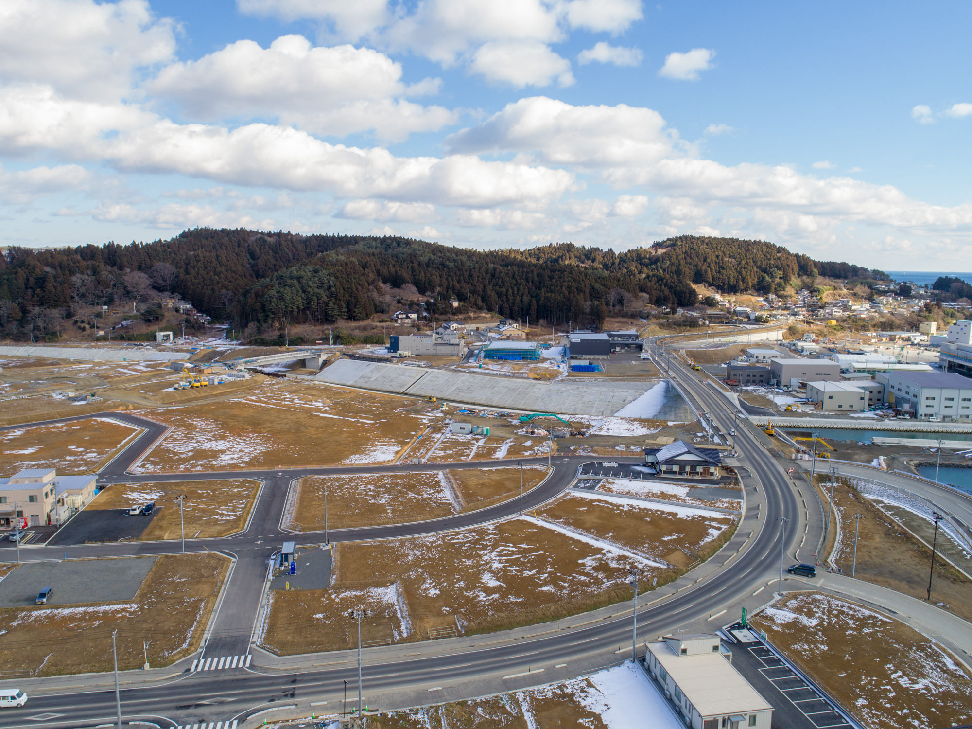 地面には雪が残っている開けた土地と道路、建物が混在する志津川地区を上空から撮影したもので、曲がりくねった道路や住宅・商業施設と思われる建物が点在し、背景には森林に覆われた丘が広がっている冬景色の写真