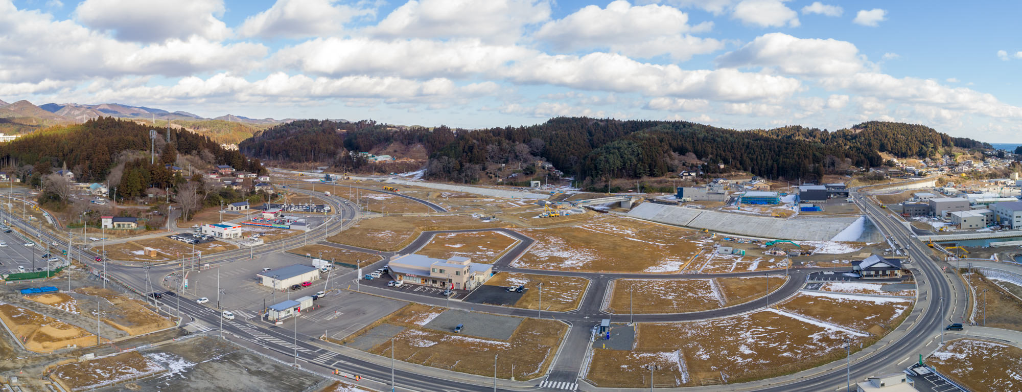 地面には雪が残っている道路や建物が広がる志津川地区を広く見渡したもので、住宅や商業施設などの構造物が点在し、周囲には木々に覆われた丘が広がり、背景には山々が見える冬景色のパノラマ写真