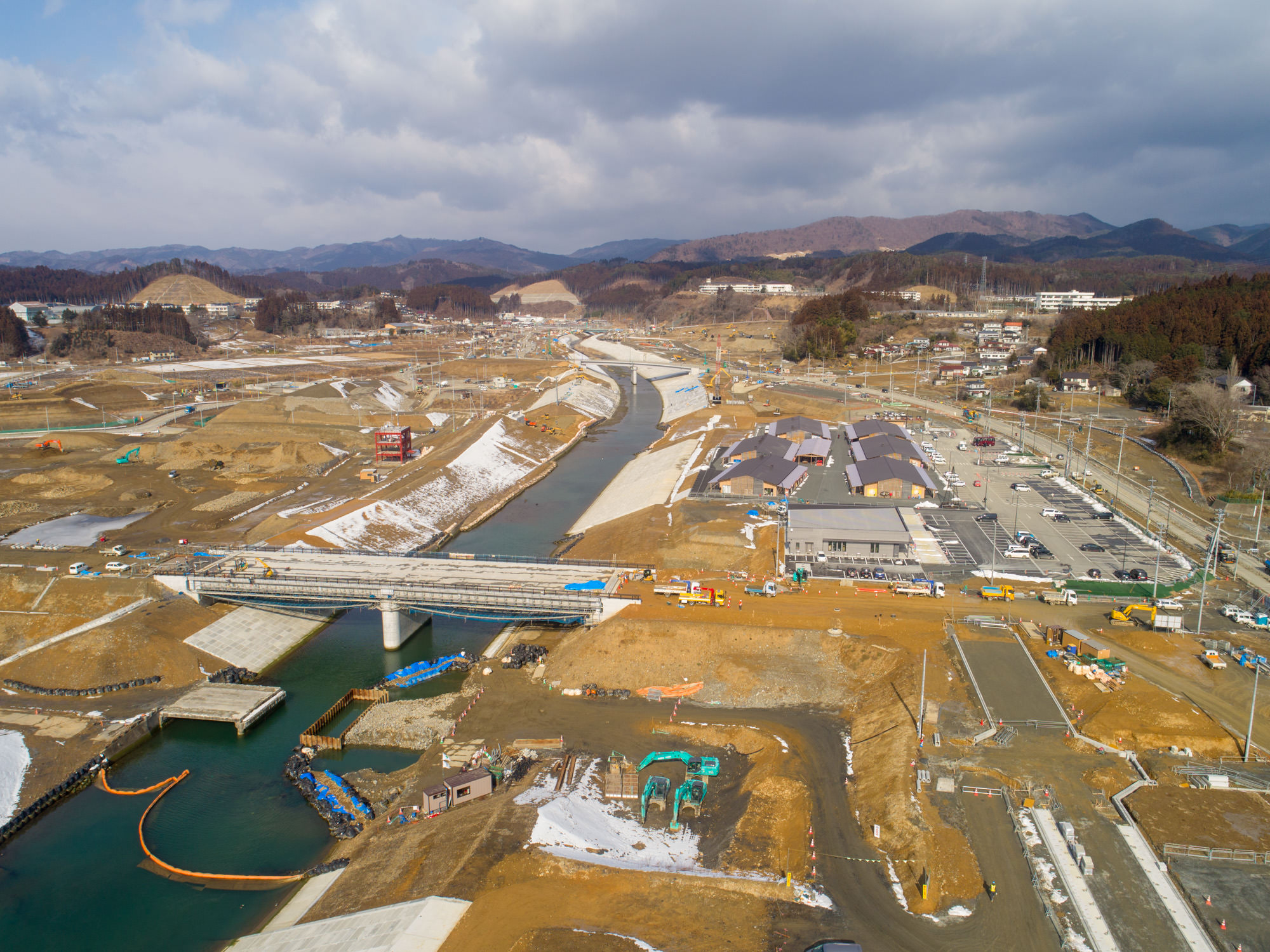 川が中央を流れる工事現場を上空から撮影した写真で、複数の橋が川を横断し、現場内には建設機材や資材が点在し、周囲には建物や駐車場、道路が整備され、背景には丘や山が広がり、地面には一部雪が残る風景の写真