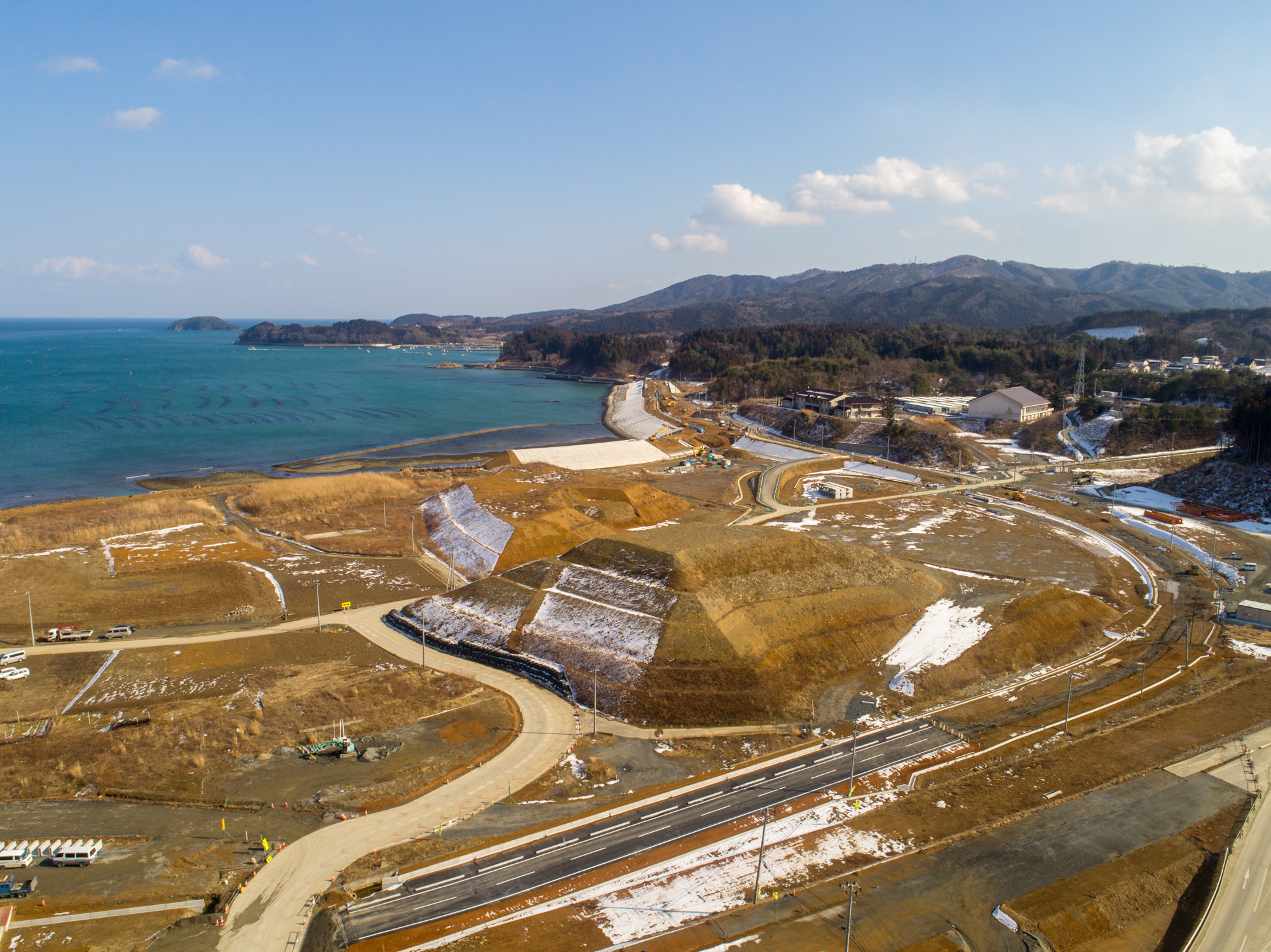 自然と人工の要素が混在する海沿いの風景を撮影した写真で、曲がりくねった道路や工事中の場所が点在し、地面には雪が残る部分もあり、左側には海岸線と海が広がり、背景には丘や山が連なり、空は晴れて雲が少し浮かぶ風景の写真