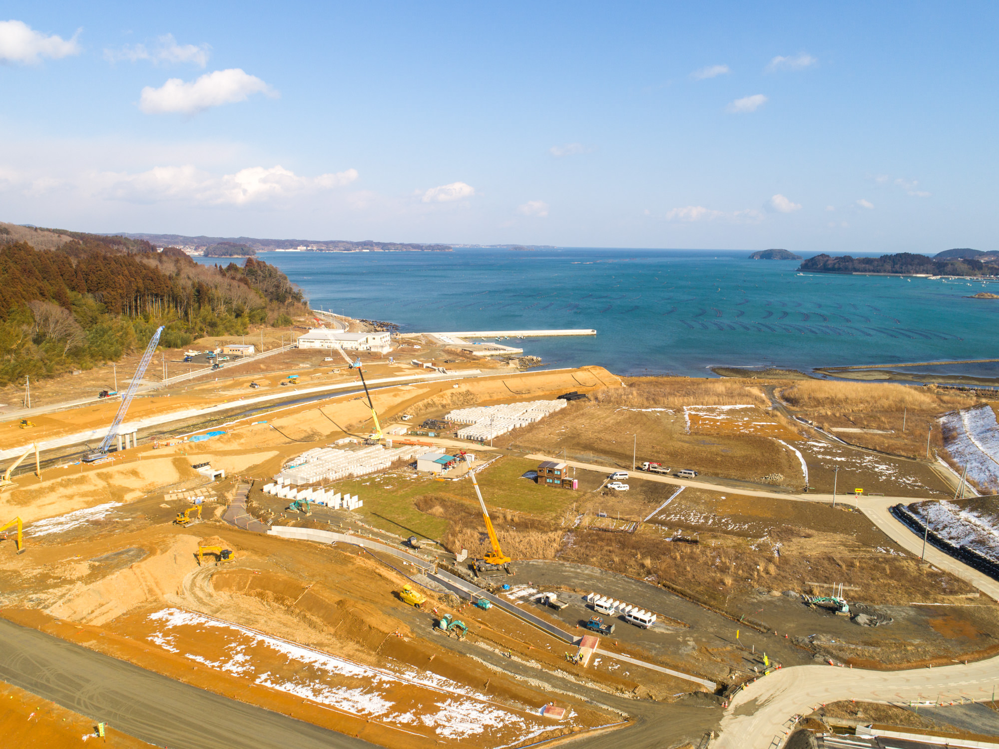 海沿いの工事現場を撮影した写真で、クレーンやトラックなどの建設車両が稼働しており、現場には建設中または完成済みの構造物が点在し、周囲の地面には雪が残っている、海と遠くの陸地が見渡せる風景の写真