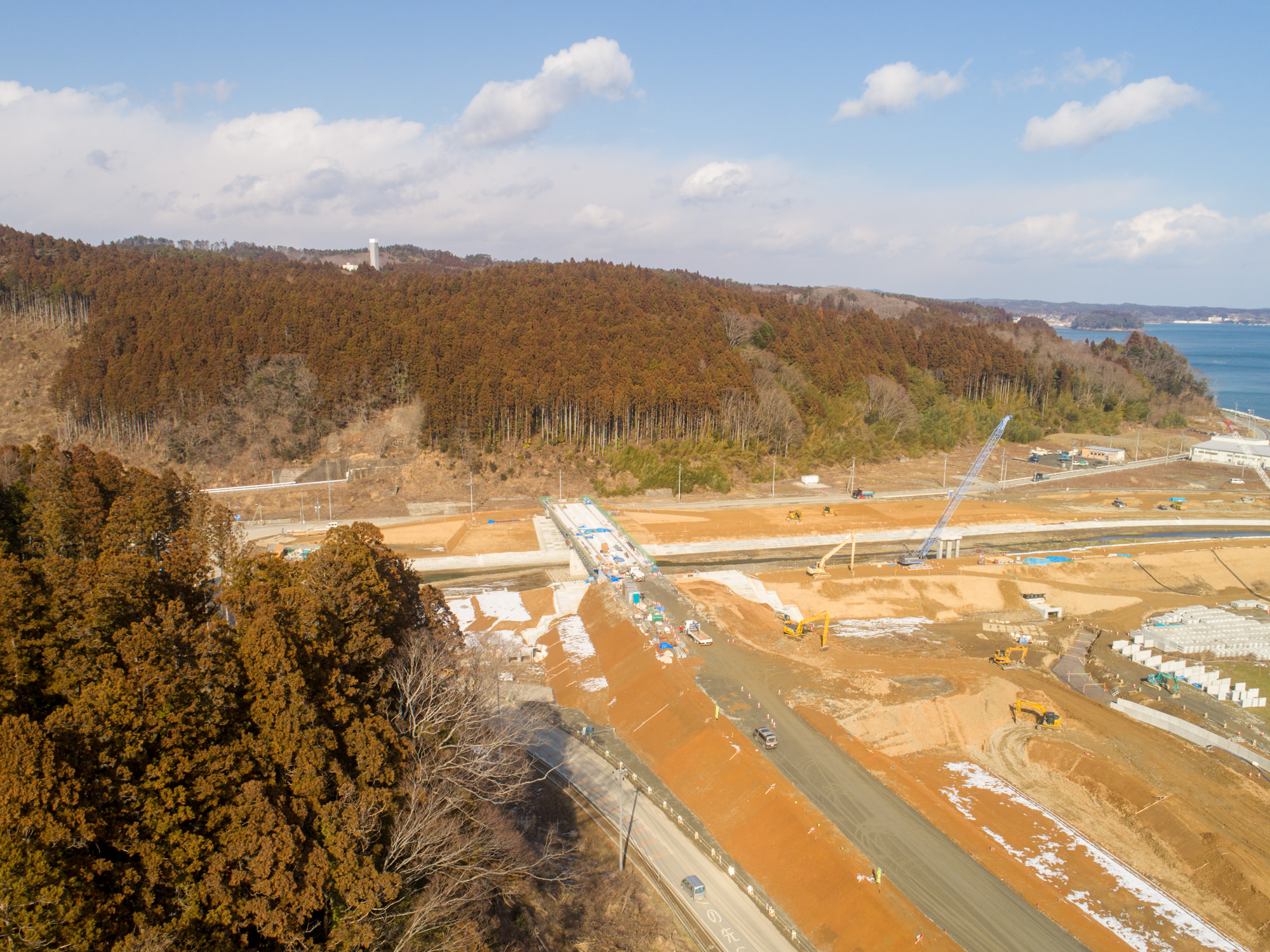 残雪のある工事中の現場を撮影した写真で、道路の建設が進められており、作業車両や作業員が見られ、背景には森林に覆われた丘と右側に海が広がり、空は雲が少し浮かぶ晴れ間のある風景の写真