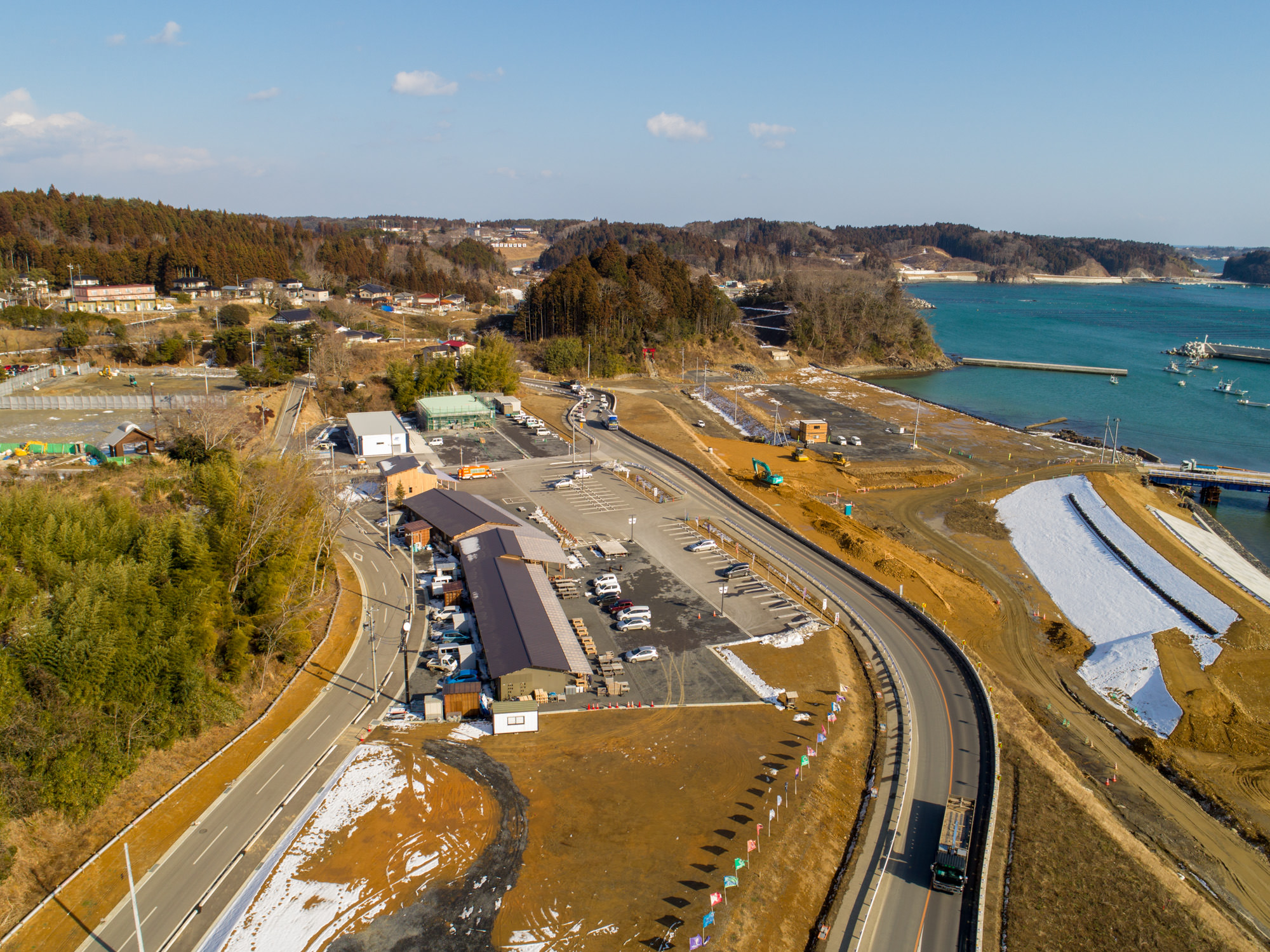 海沿いの地域を上空から撮影したもので、湾曲した道路が駐車場や複数の建物の周囲を通り、駐車場には車両が停まっており、水辺には建設機械も見られ、背景には木々や丘が広がる自然と開発が混在した風景が広がっている海沿いの街と建設風景の写真