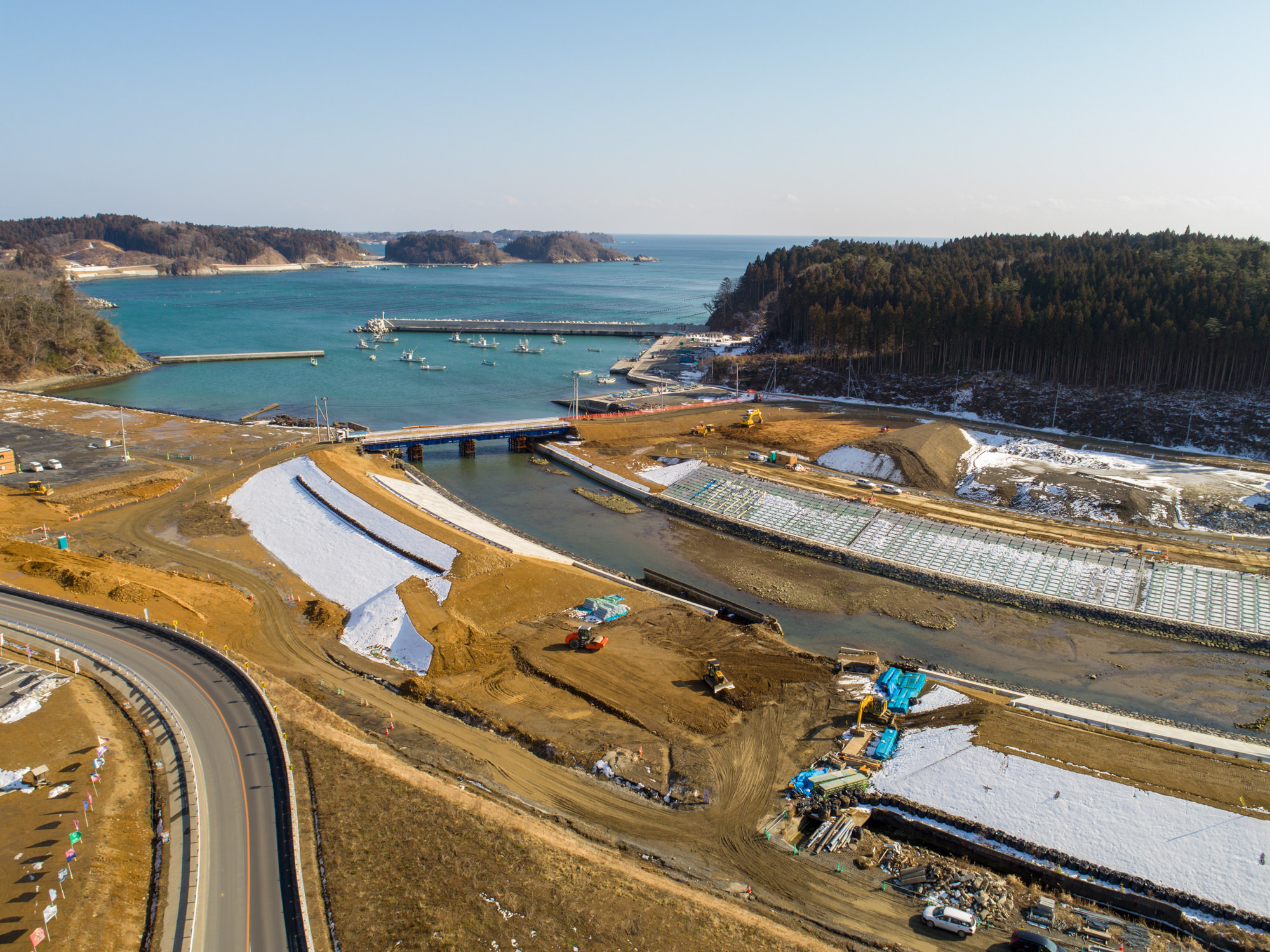 海に面した建設現場を写したもので、手前には湾曲した道路が通り、工事が進行中のエリアには白いシートで覆われた地面や建設資材・機械が見られ、水路に架かる橋の先には港に停泊する複数の船があり、背景には森林に覆われた丘や遠くの島々が広がっている海辺の建設現場と港の写真