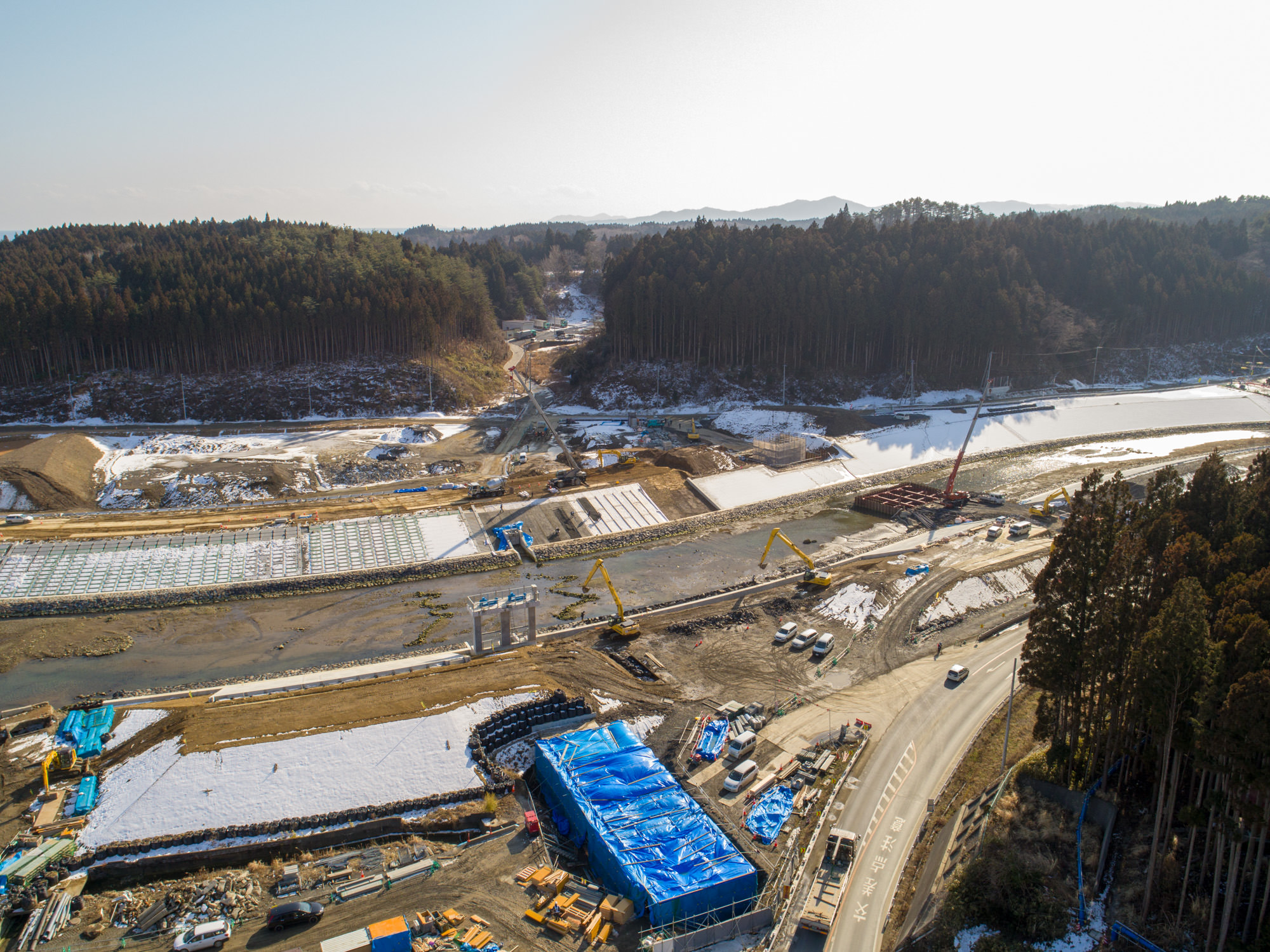 森林に囲まれた地方の建設現場を写したもので、クレーンや車両、大きな青いシートで覆われた構造物などの建設資材・機械が点在し、近くには川や水辺があり、地面には雪が残っている様子が見られる森林地帯の建設現場の写真