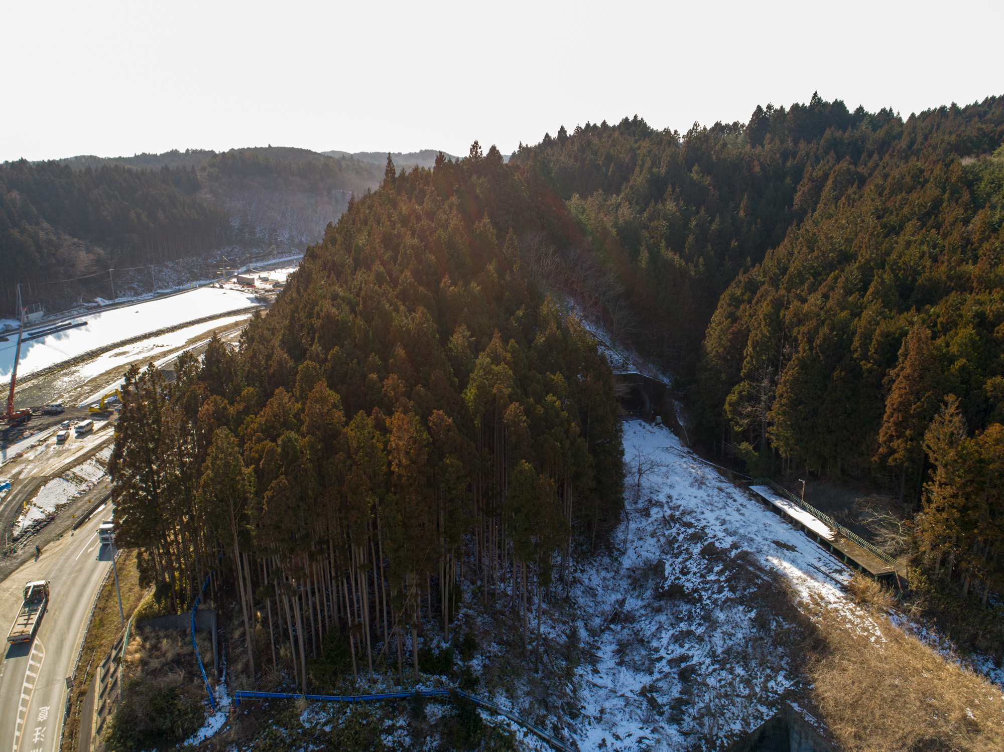 針葉樹が広がる森林地帯を写したもので、太陽の日差しが当たらない木の陰になっている地面には雪が残り、左側には道路が通り、その近くに建設機械が見られ、丘には木々が密集しており、丘の一部にはトンネルや通路のような構造が確認できる森林と道路、トンネルのある風景の写真