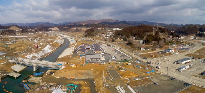 志津川地区の工事現場を上空から撮影した写真で、複数の建物や道路、橋が建設中であり、中央には川が流れ、いくつかの橋がその上を横断し、背景には雪をかぶった山や丘が広がり、現場には建設機材や資材が点在する工事現場の写真