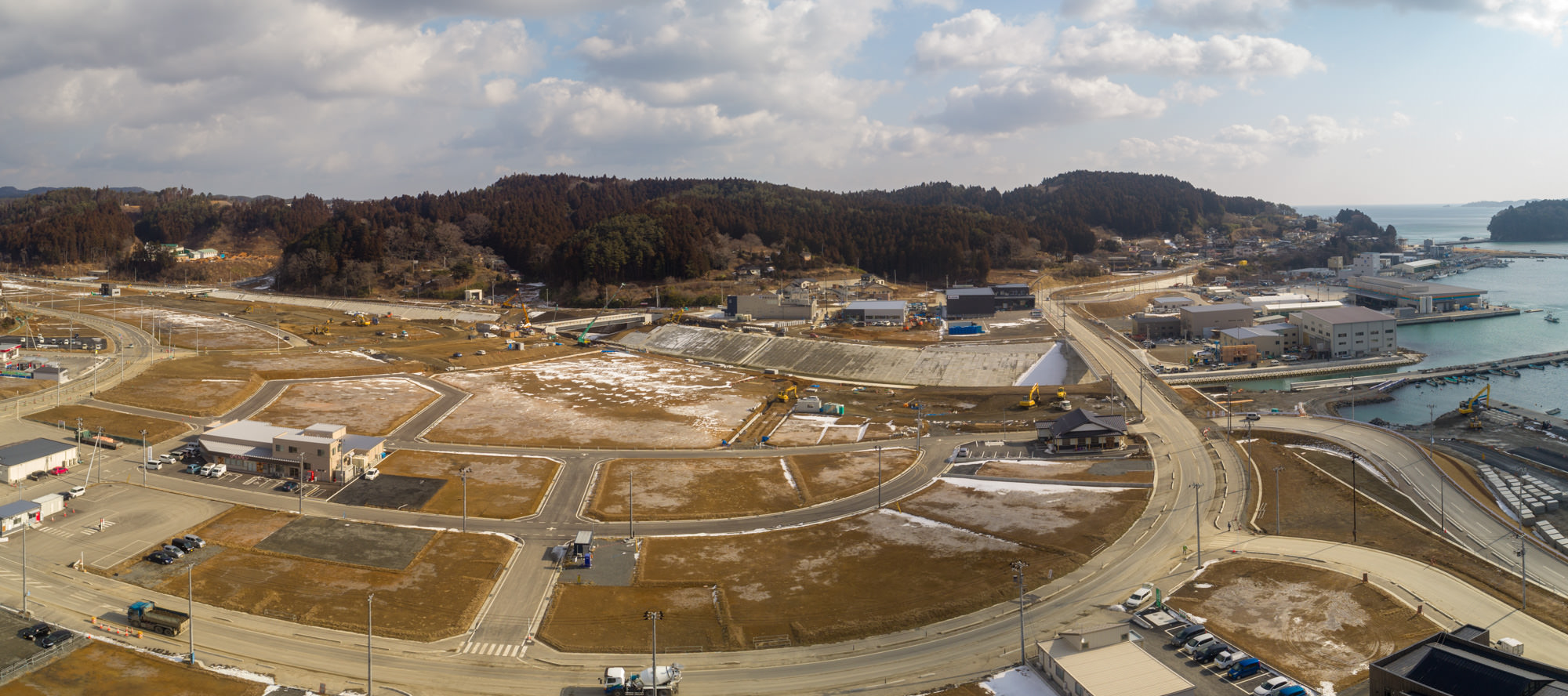 海沿いの町を上空から撮影した写真で、複数の道路や建物、空き地が広がり、地面には一部雪が残っていて、重機やトラックが稼働していて、ところどころ工事中の箇所があり、右側には海岸線と水面が広がり、桟橋や船が見られ、背景には森林に覆われた丘と雲が少し浮かぶ空が広がる風景のパノラマ写真