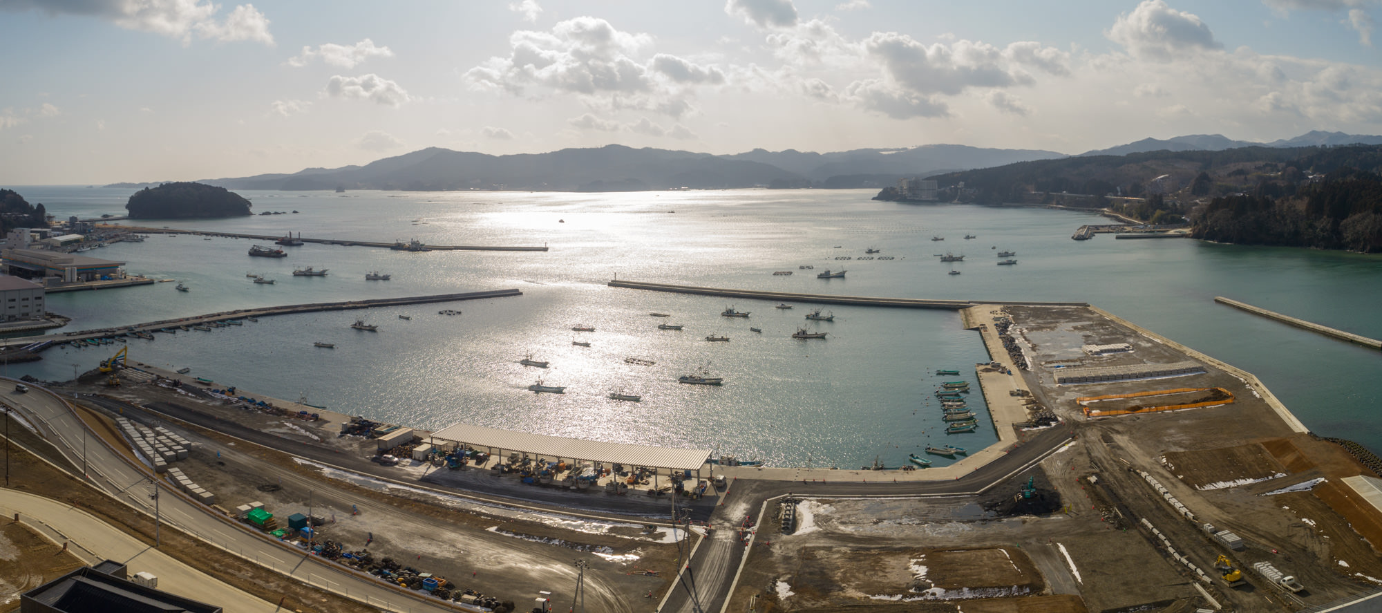 港のある海沿いの地域を広く見渡したパノラマ写真で、海には複数の船が停泊し、防波堤が港を囲み、周囲の陸地では道路や建設機材、資材が見られ開発が進められており、背景には丘や山、そして地平線まで広がる水面があり、空は雲が少し浮かび太陽の光が水面に反射する風景の写真