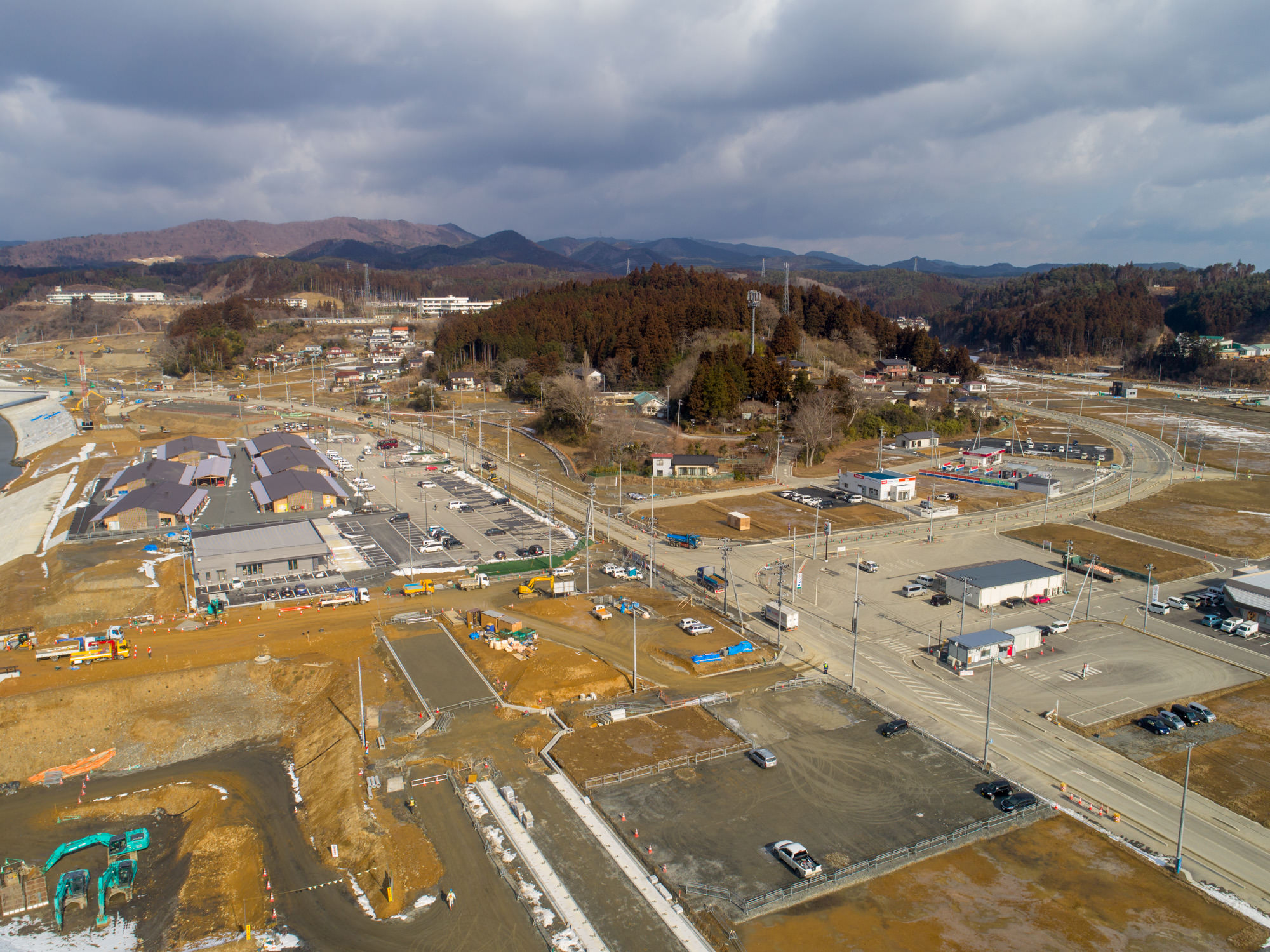 志津川地区を上空から撮影した写真で、左側には倉庫や商業施設と思われる大きな建物が並び、道路や工事現場が点在し、複数の車両や建設機材が見られ、背景には丘や山が広がり、空は曇っていてる風景の写真