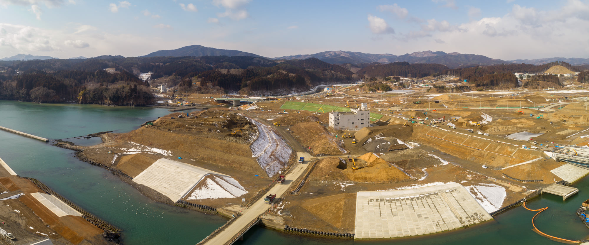 開発または再整備中の海沿いの地域を広く見渡した写真で、水辺の近くには大型のコンクリート構造物や堤防があり、一部の地面には雪が残り、背景には丘や山が広がり、現場には建物や建設機材が点在する風景のパノラマ写真