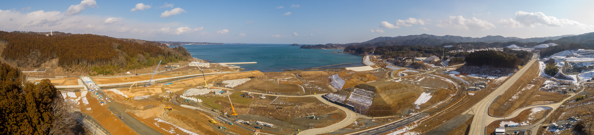 開発または工事中の海沿いの地域を広く見渡した写真で、現場にはクレーンを含むさまざまな建設車両や機材が点在し、丘や道路、雪の残る場所があり、海岸線が広がって海が地平線まで続き、背景には森林や山々が広がる風景のパノラマ写真