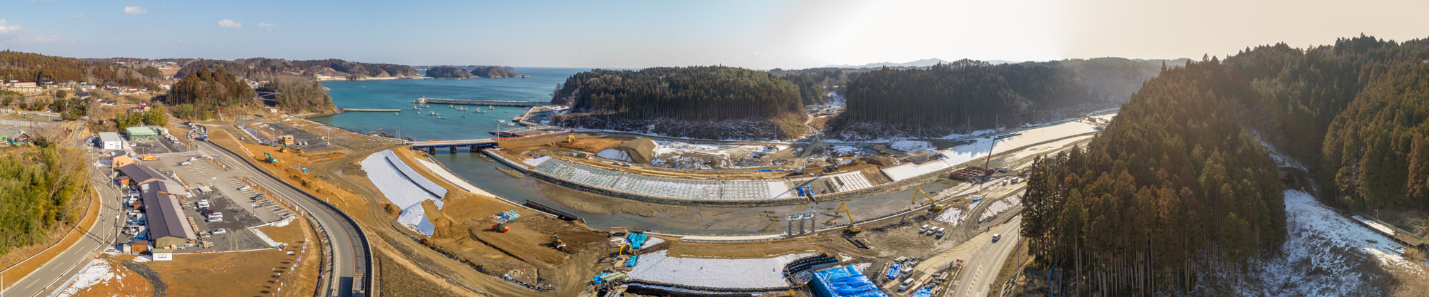 海が広がる水域を背景に、丘陵や森林に囲まれた沿岸地域を広く見渡したもので、手前には建設現場が広がり、重機や資材が点在し、左側には道路や建物も見られ、地面には雪が残る寒冷地の様子が写されている沿岸地域の建設風景の写真