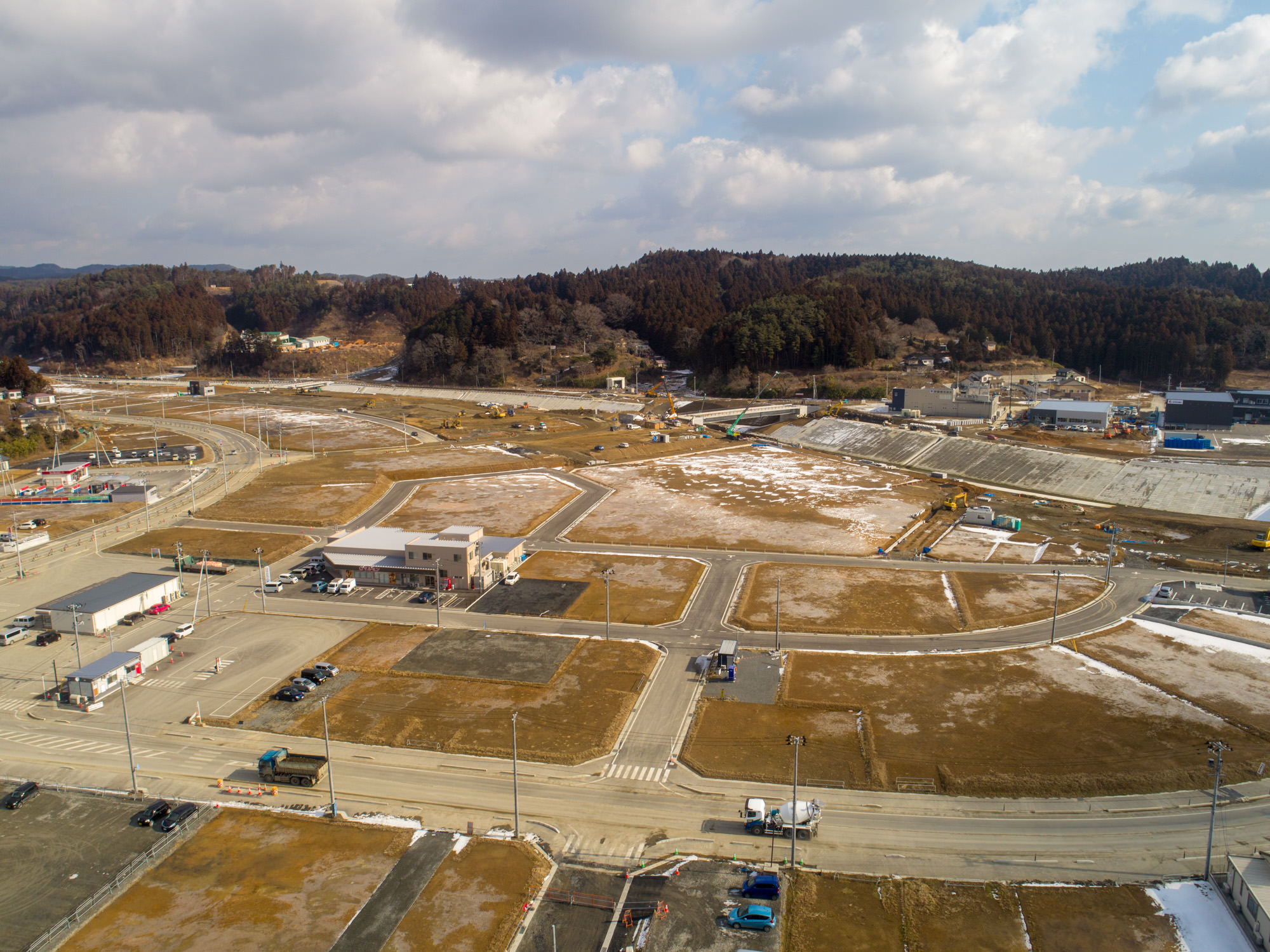 海沿いの町を上空から撮影した写真で、複数の道路や建物、空き地が広がり、地面には一部雪が残っていて、工事中の地域と思われ、右側には海岸線と水面が広がり、桟橋や船が見られ、背景には森林に覆われた丘と雲が少し浮かぶ空が広がる風景の写真